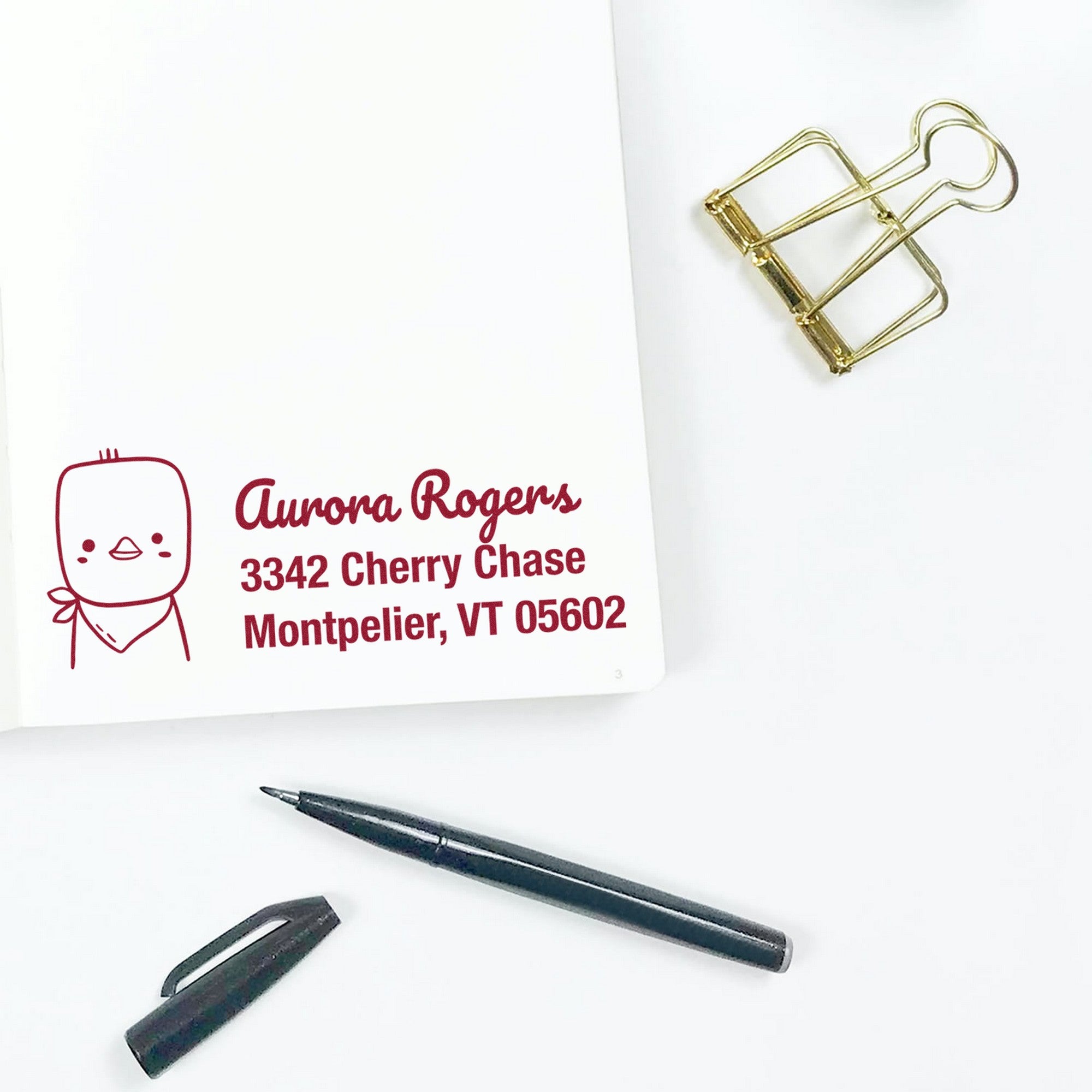 Wood handle cute chick handmade mail address rubber stamp on white paper, displaying customizable return mail stamper with name and address in red ink. Nearby are a black pen, cap, and gold binder clip on a white surface.