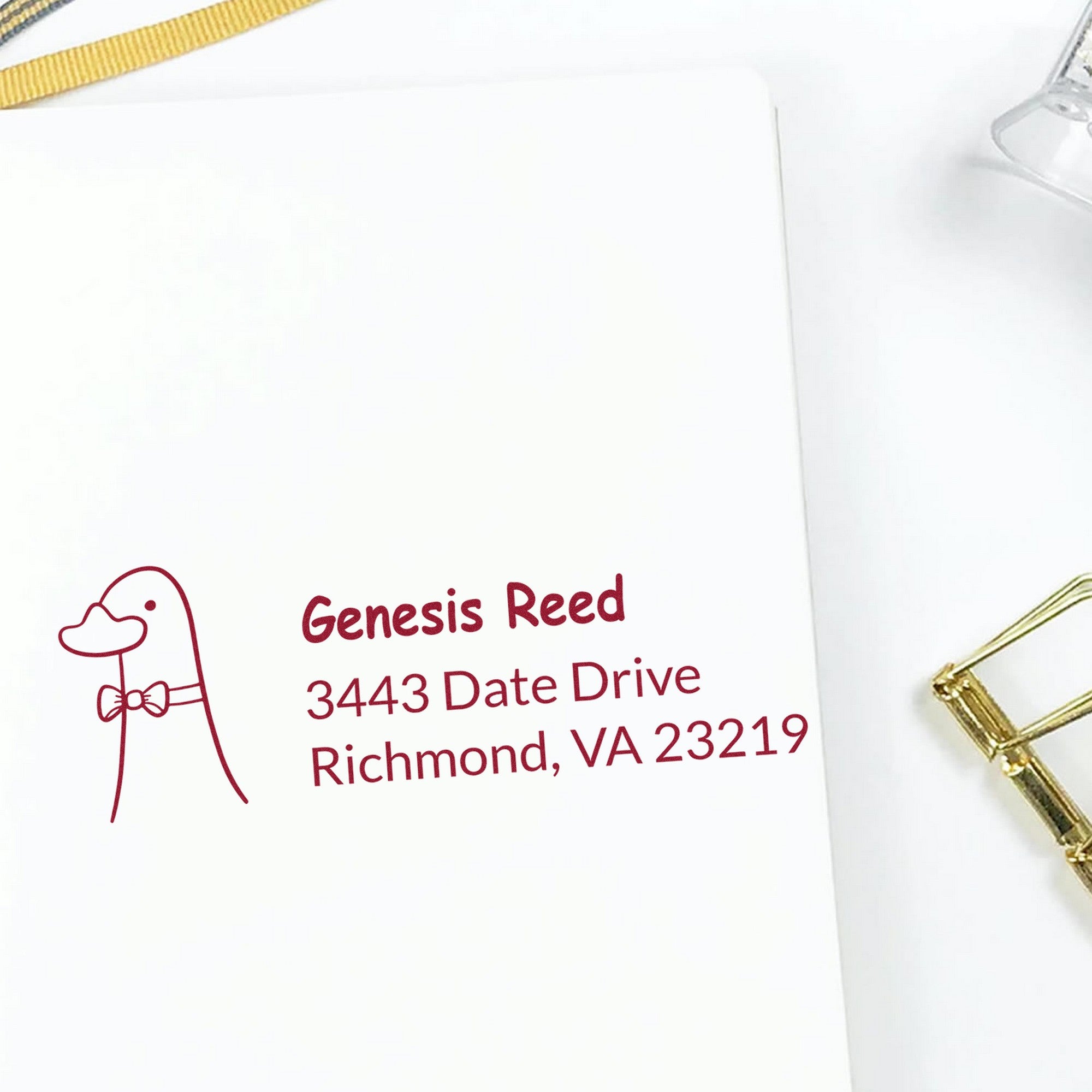 Dashing Duck Customized Address Self-Inking Stamp displayed on white paper, featuring a duck illustration and text: "Genesis Reed, 3443 Date Drive, Richmond, VA 23219." This Customized Return Mail Stamper is shown with a gold paperclip nearby.