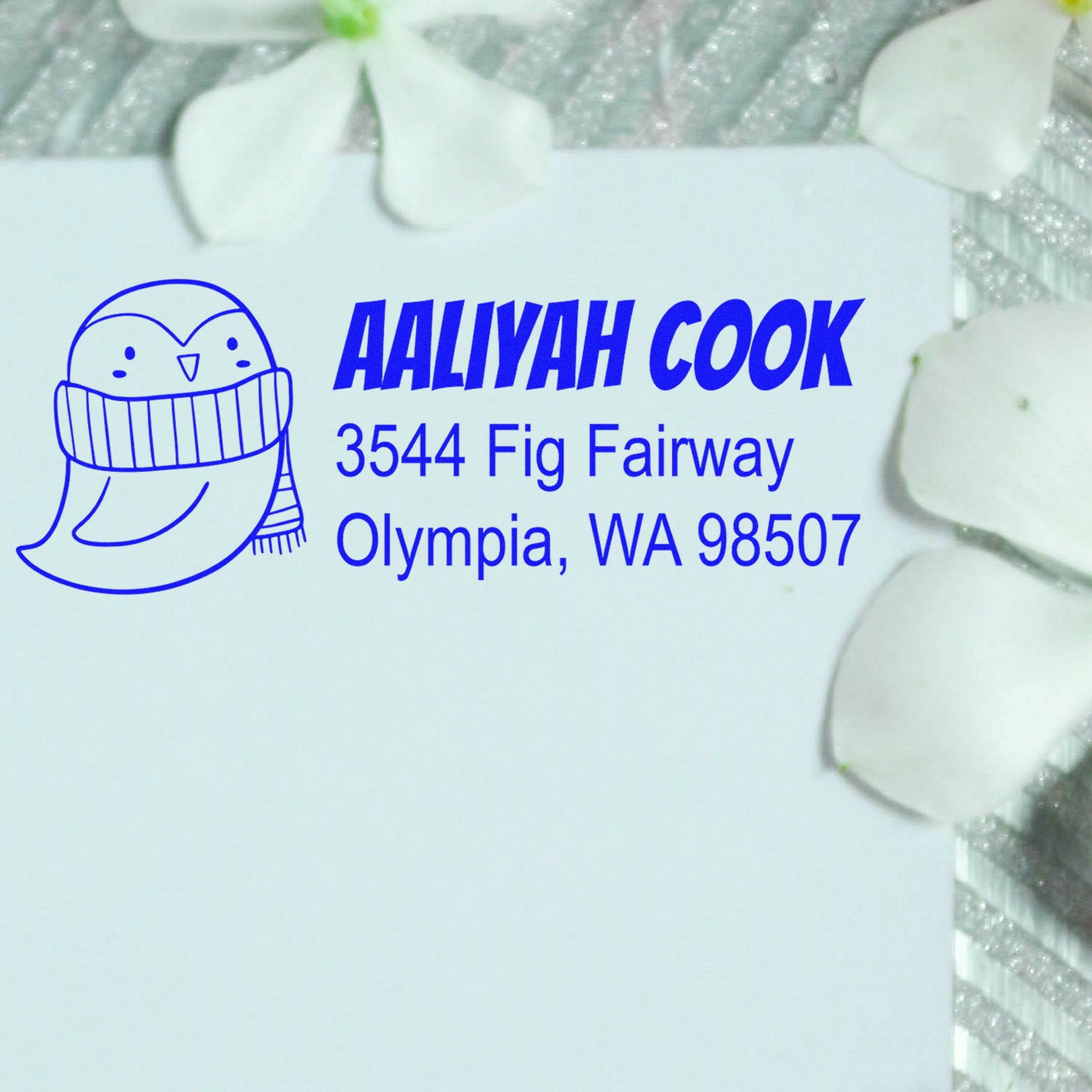 Pretty Penguin Customized Return Address Self-Inking Stamp on an envelope, featuring a cute penguin illustration. The Custom-Made Return Mail Stamp displays the name "Aaliyah Cook" with an address in Olympia, WA, surrounded by white flower petals.
