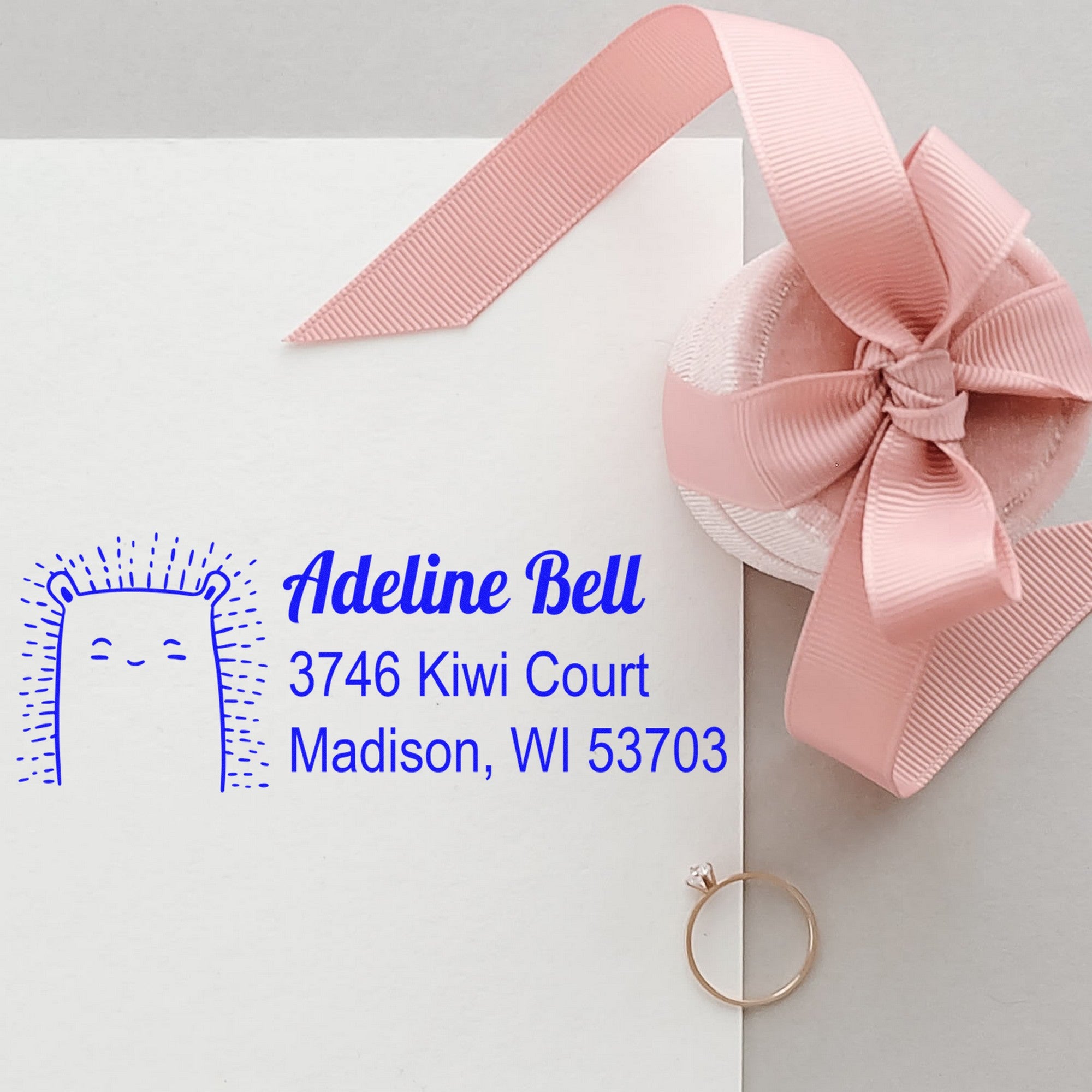PSI Bouncy Bear Personalized Address Return Pre-Inked Stamp on an envelope with "Adeline Bell, 3746 Kiwi Court, Madison, WI 53703" in blue. A pink ribbon and ring are nearby. Personalizable Mailing Stamp for custom correspondence.