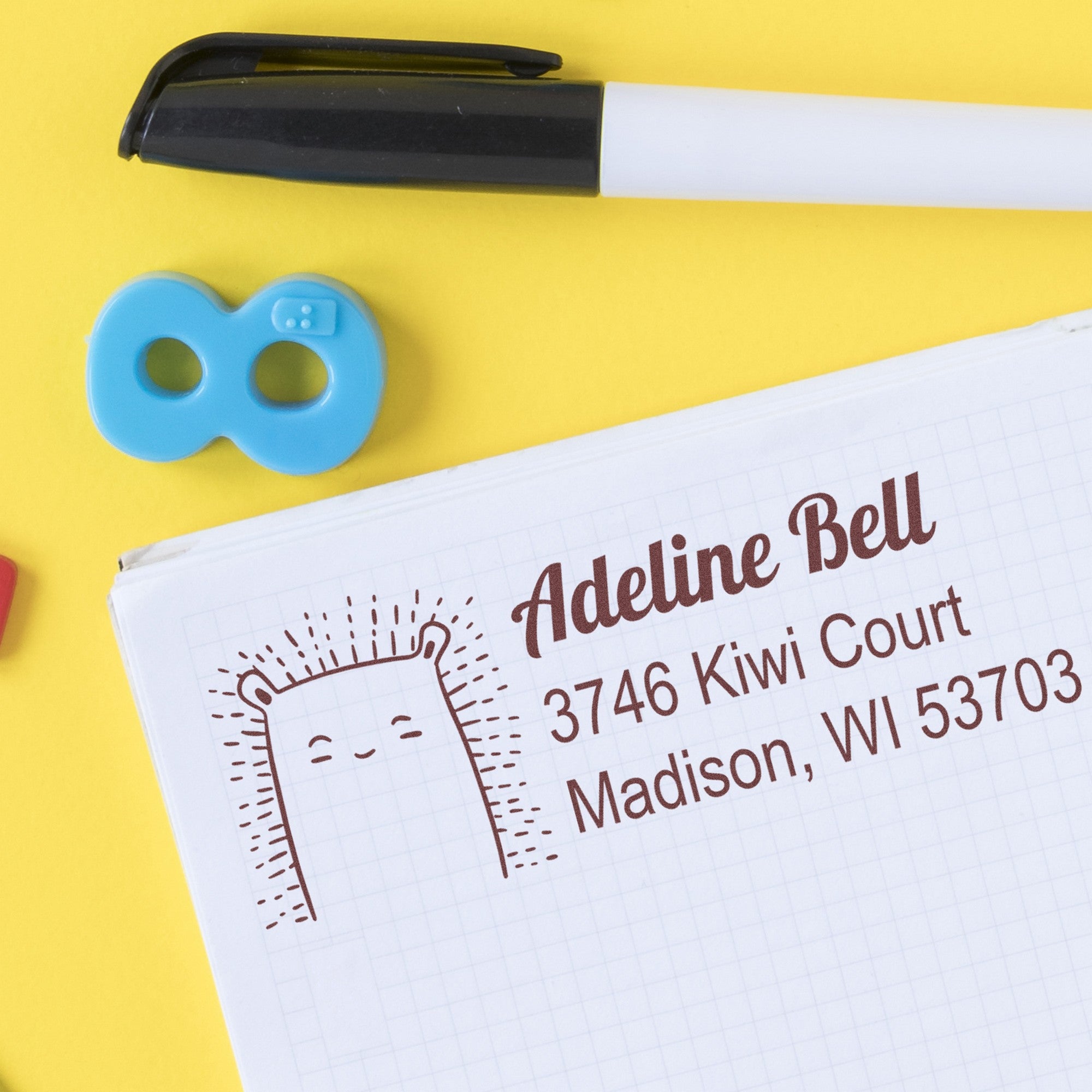 PSI Bouncy Bear Personalized Address Return Pre-Inked Stamp on a notebook page with "Adeline Bell, 3746 Kiwi Court, Madison, WI 53703" in brown ink. A pen and a blue paperclip are nearby. Personalizable Home Stamp for custom addresses.