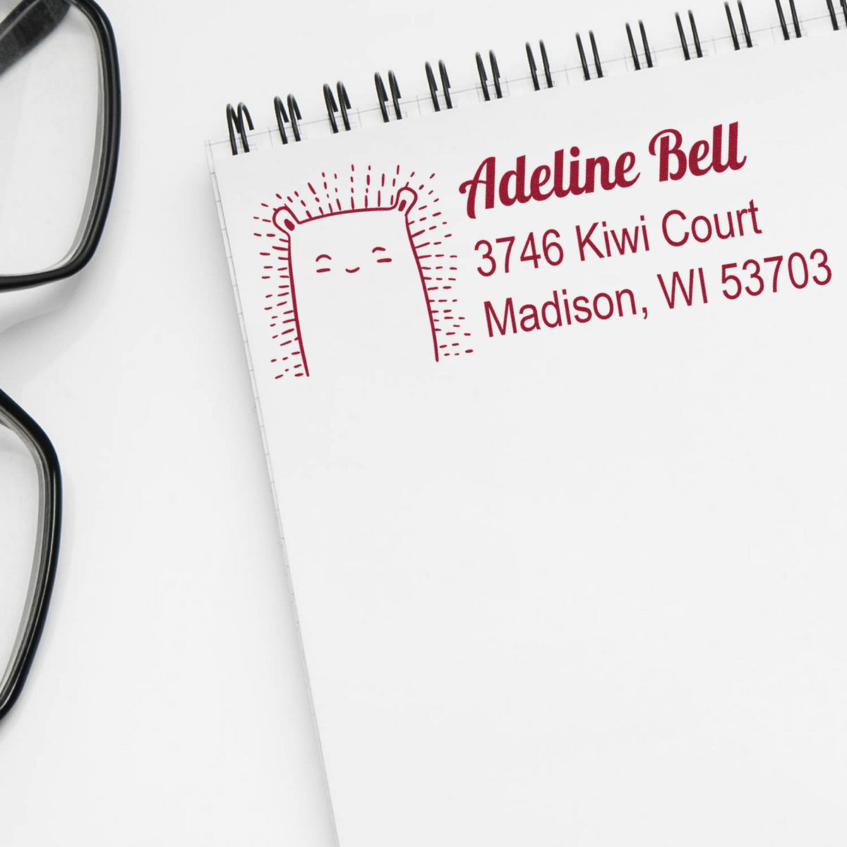 A notebook page displays a stamped image of a smiling bear with the text Adeline Bell, 3746 Kiwi Court, Madison, WI 53703 in red. The Slim Pre-Inked Bouncy Bear Customize New Address Stamp is used as a Personalized Return Mail Stamp. Glasses are nearby.