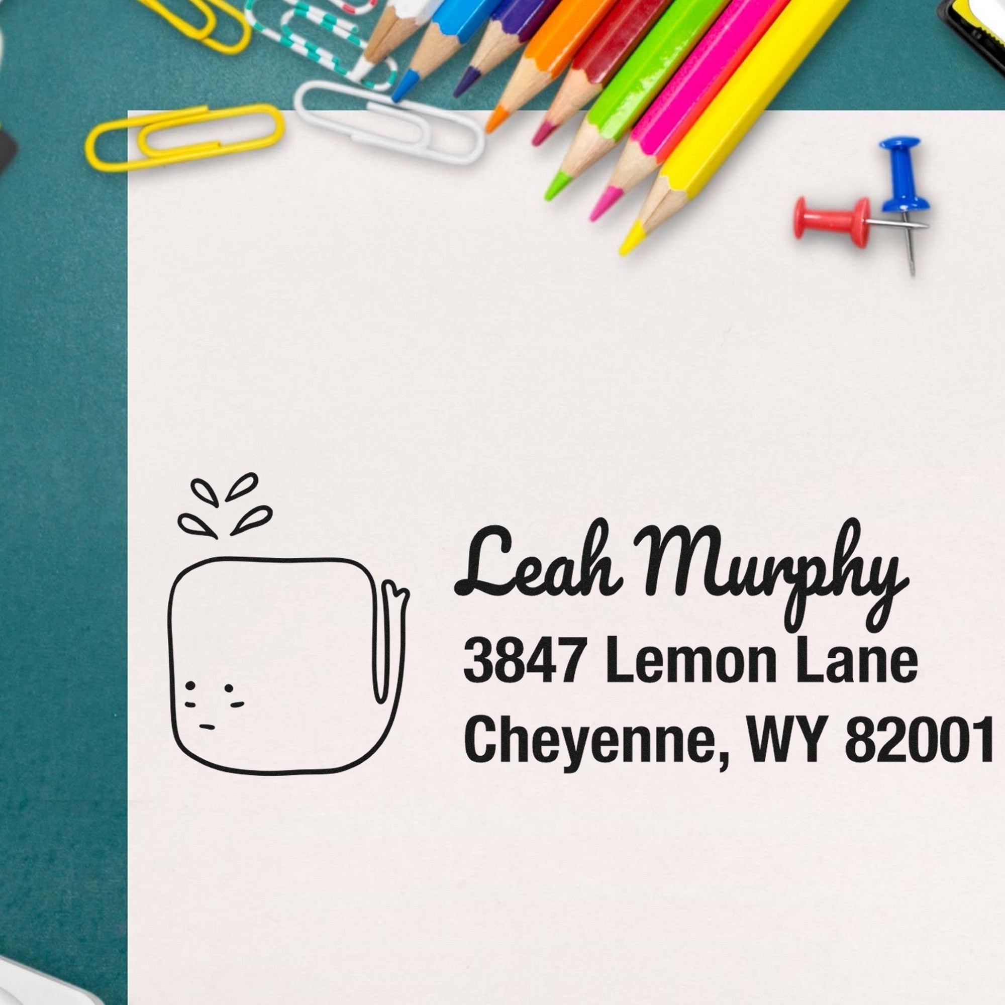 Wood handle joyful whale customize return address rubber stamp on paper with "Leah Murphy, 3847 Lemon Lane, Cheyenne, WY 82001" text. Surrounding are colorful pencils, paperclips, and push pins. Custom mailing stamp design.