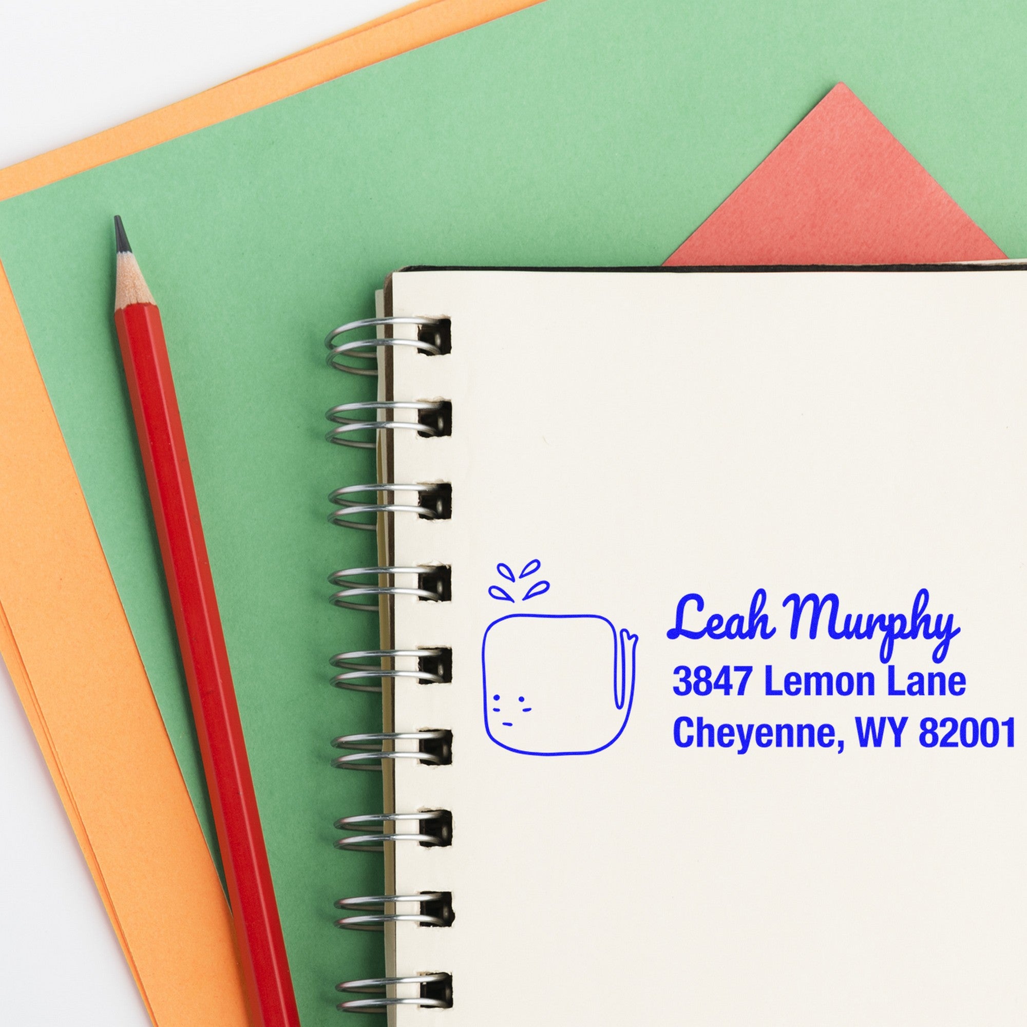 A notebook with a red pencil on a green and orange background features a blue imprint from the Joyful Whale Customized Mail Self-Inking Stamp. The custom stamp displays a whale design with the name Leah Murphy and an address in Cheyenne, WY.