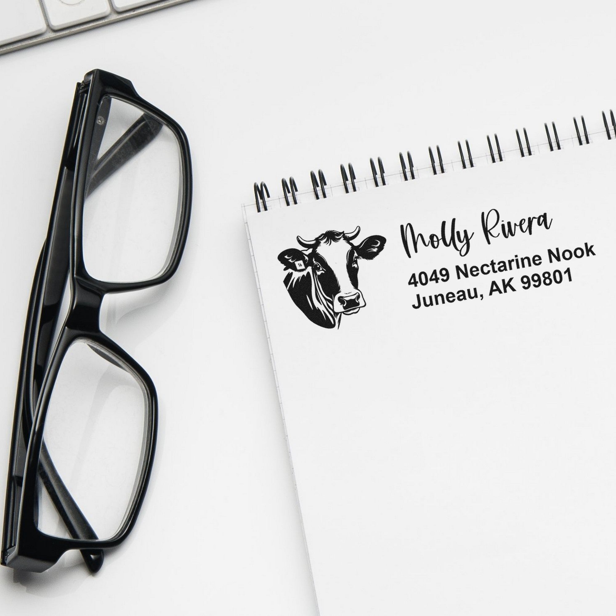 Black glasses beside a white notebook with a custom cow return-address stamp and seal on a clean white background.