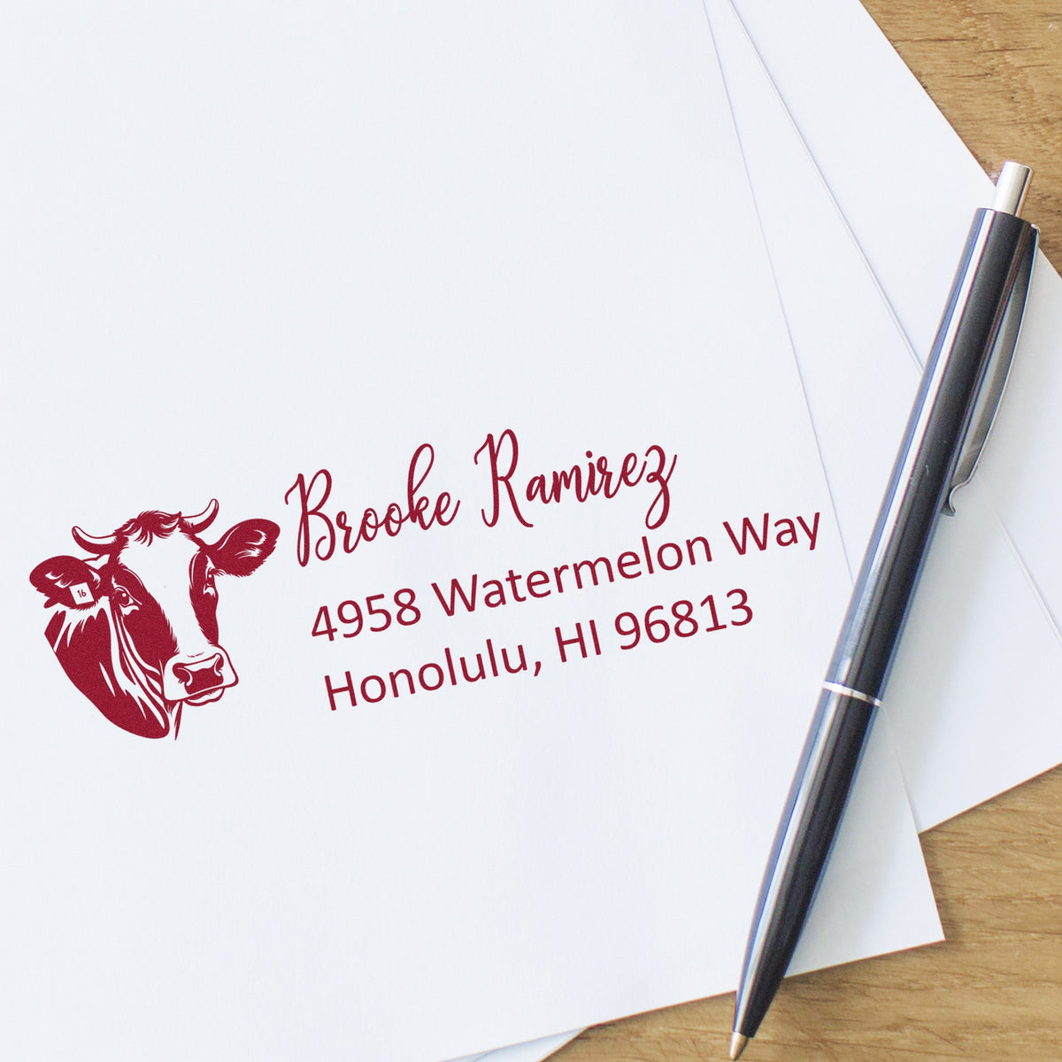 Red custom stamp and seal with cow logo and address on white envelopes, beside a black pen on a wood desk.