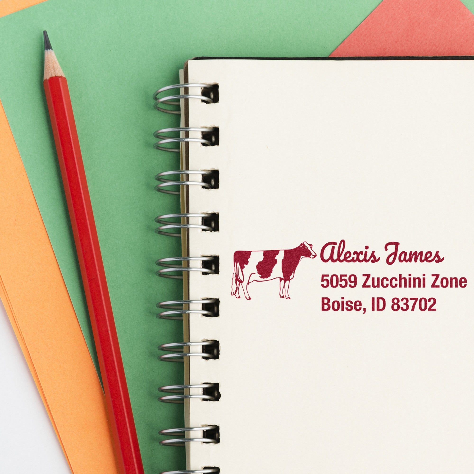 Custom burgundy cow stamp and seal on white spiral notebook beside red pencil, with green and orange paper.