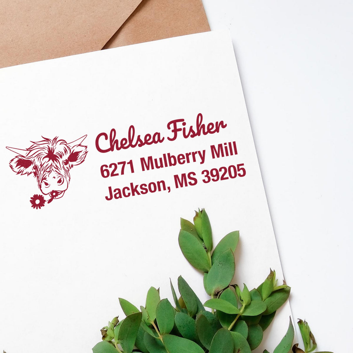 Burgundy custom stamp and seal with cow head graphic and address on a white envelope, beside kraft paper and greenery.