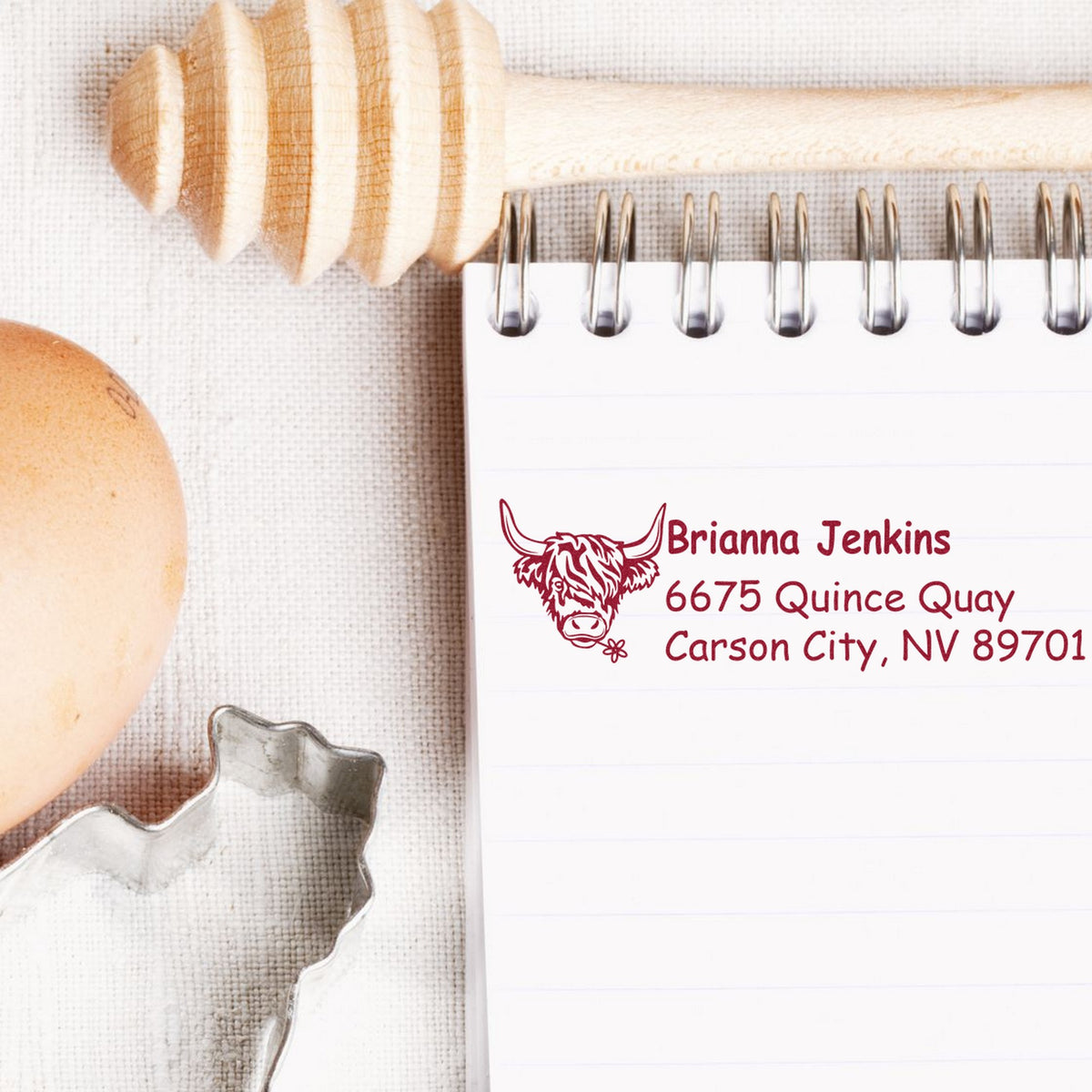 Burgundy custom cow address stamp on white notepad, beside a rolling pin, egg, and cookie cutters.