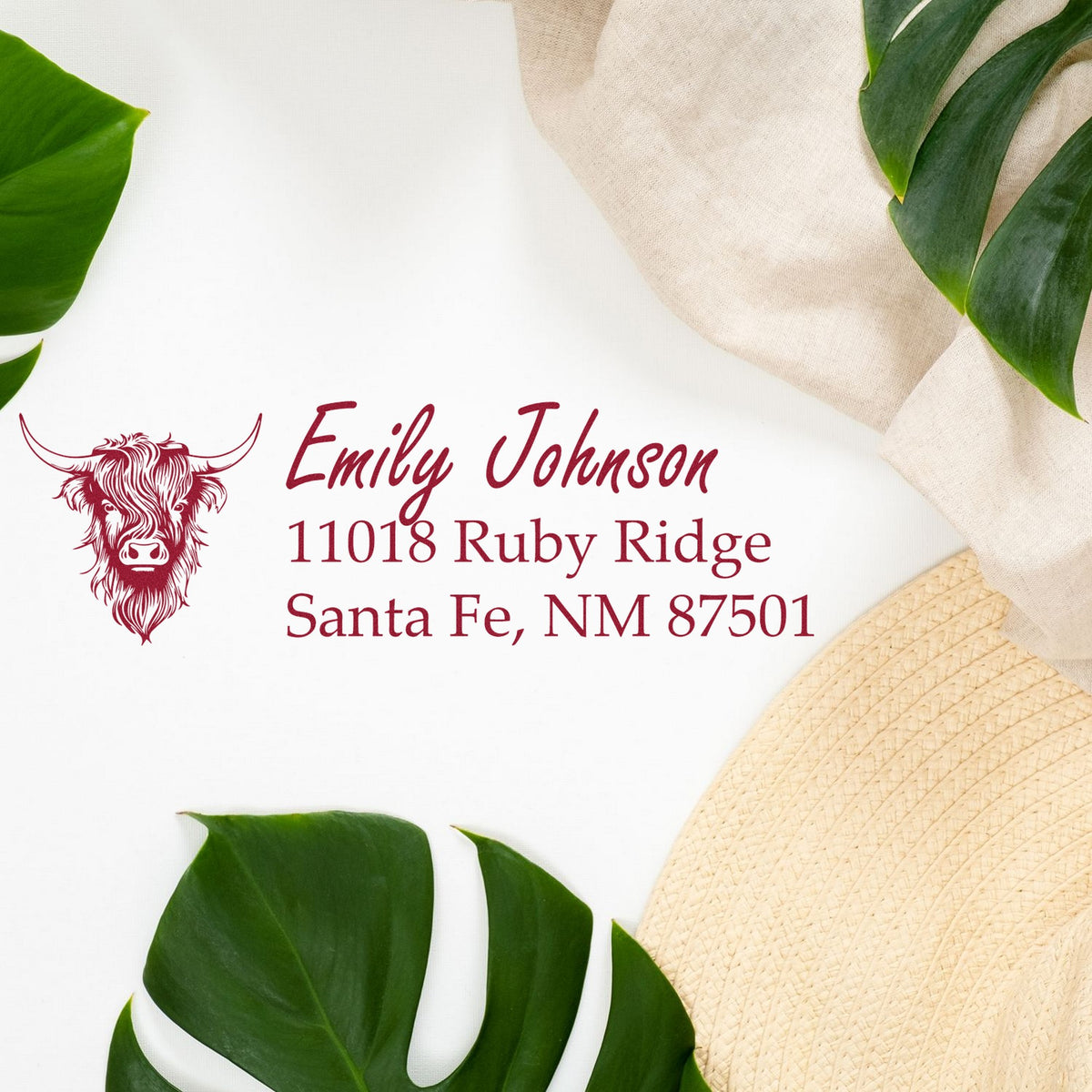 Burgundy custom return address stamp and cow seal with Emily Johnson, on white background with green leaves and beige fabric.