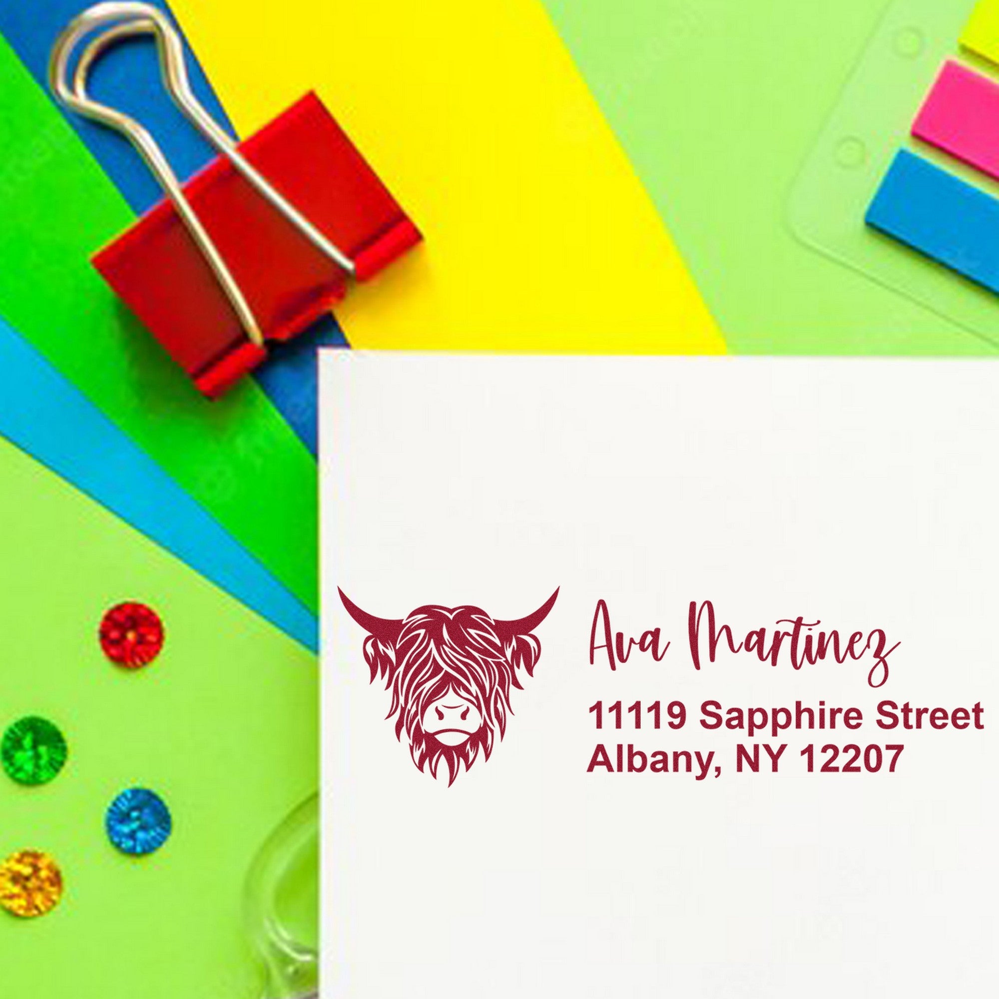 Custom burgundy stamp and seal on white envelope with bull logo, name and address, set against bright rainbow paperclips.