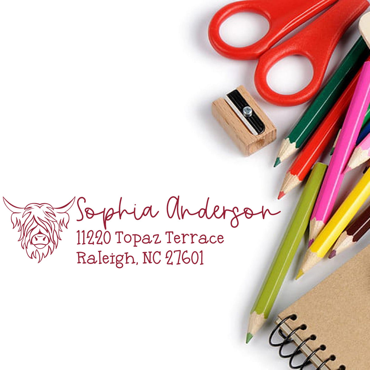 Custom burgundy address stamp and seal with lion logo, red scissors, colored pencils, and notebook on white background.
