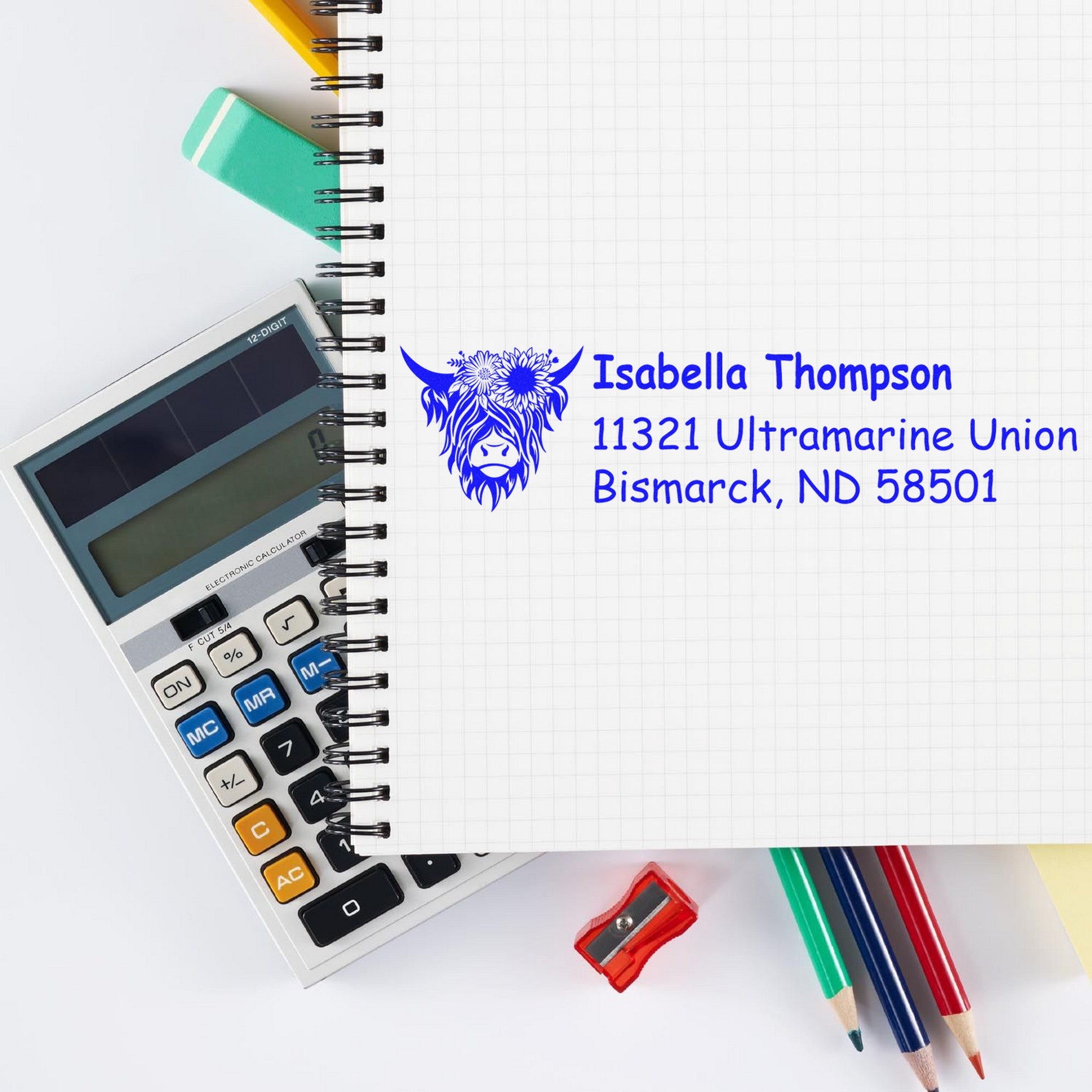 Blue custom business stamp seal with cow logo, name and address on a graph notebook, beside calculator, pencils and sharpener.