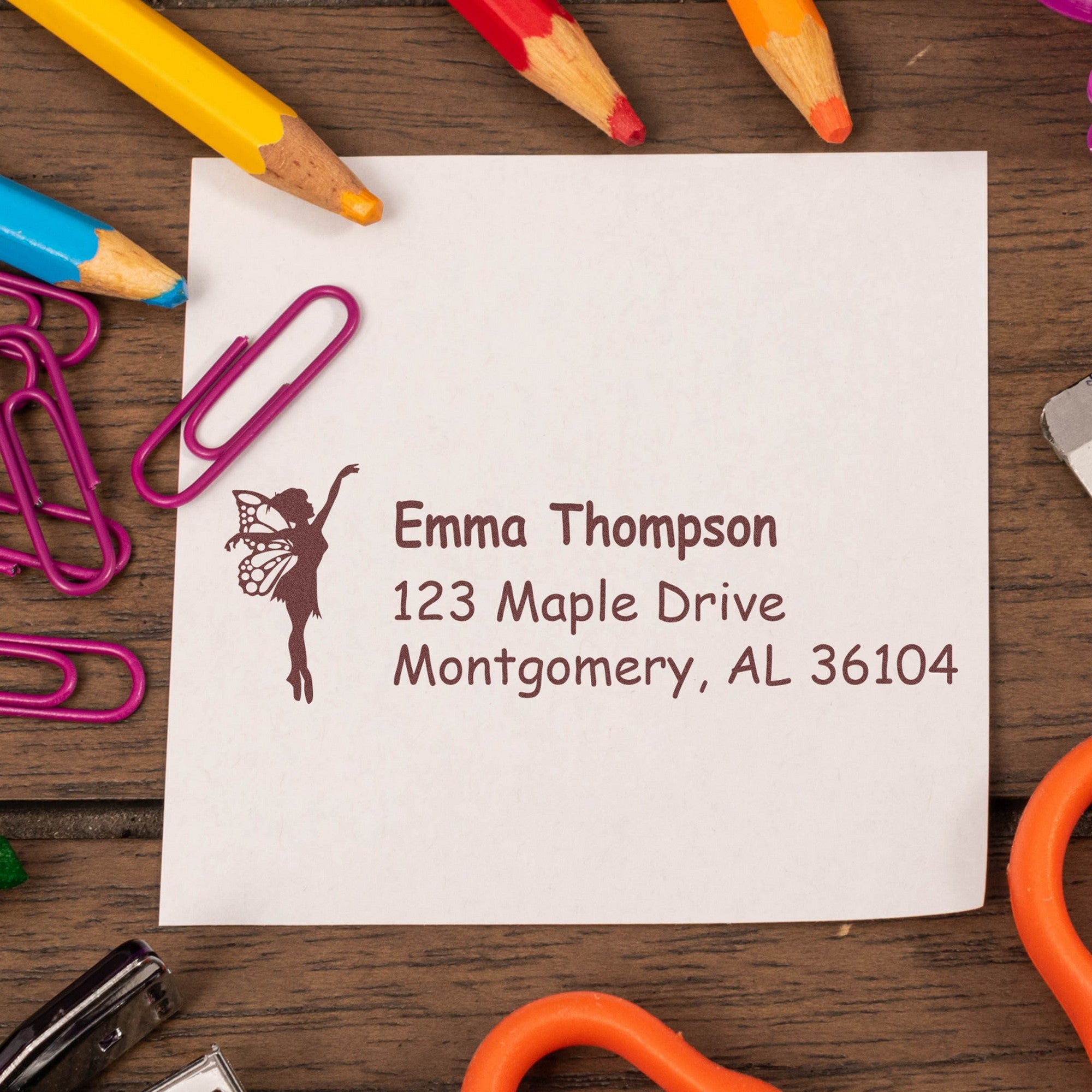 Custom brown address stamp and seal with fairy silhouette on white paper, surrounded by colorful pencils and magenta paper clips.
