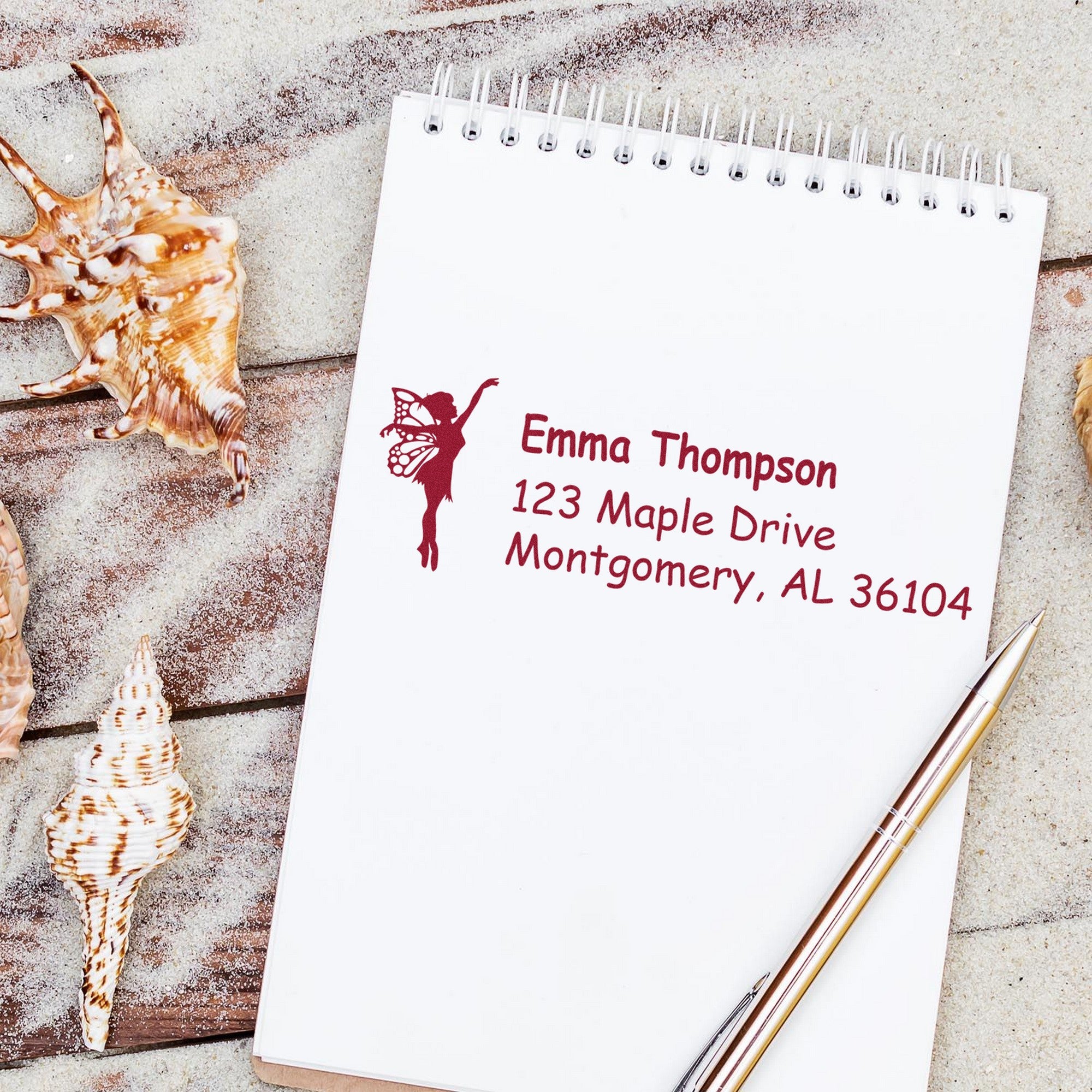 Burgundy custom address stamp on white spiral notepad, with fairy silhouette, seashells, and pens on a sandy background.