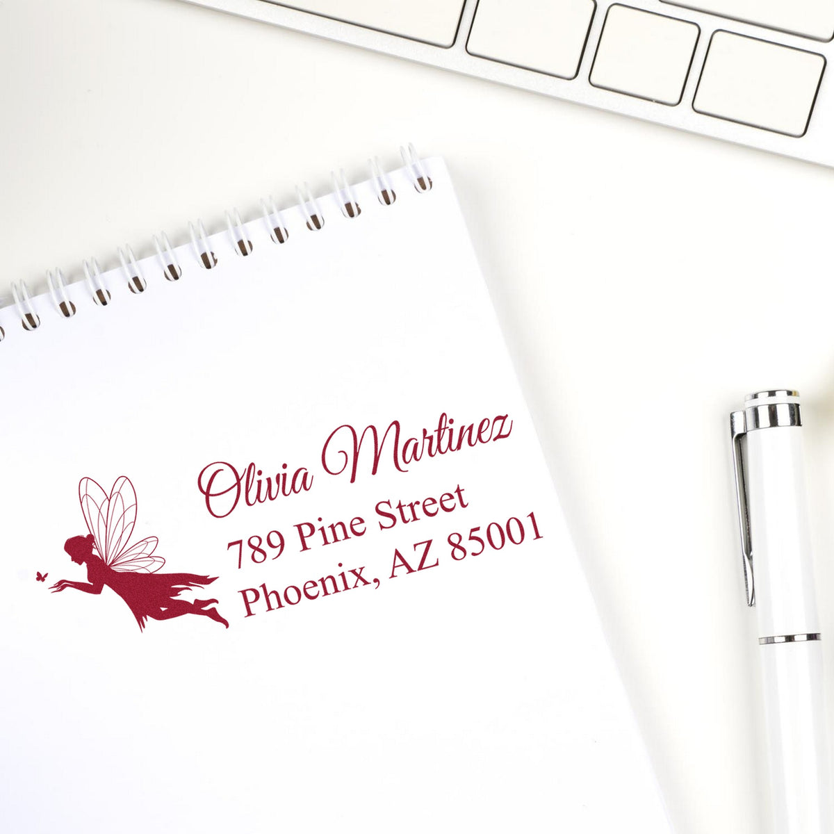 Burgundy custom address stamp and fairy seal on white notepad, beside a silver pen and keyboard.