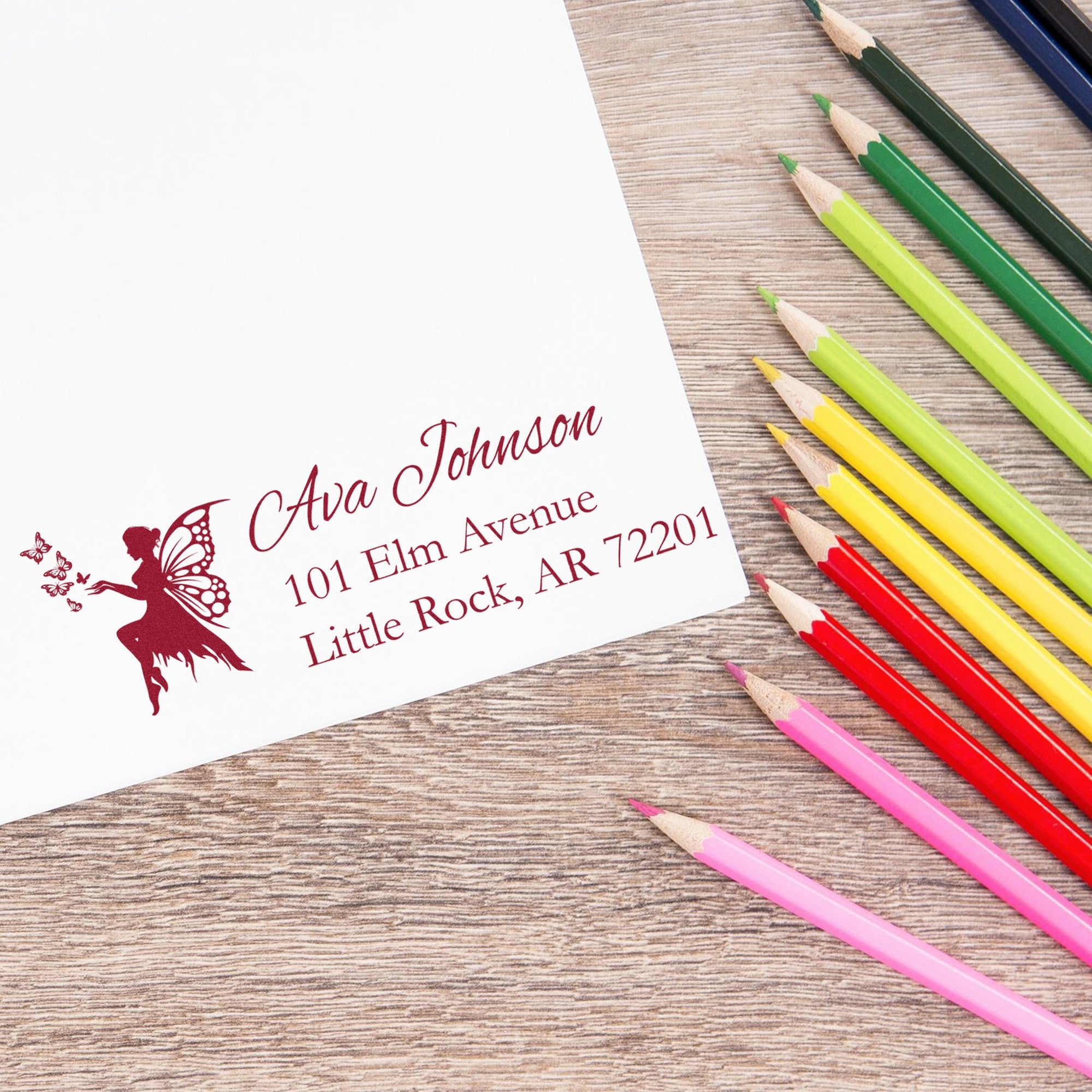 Custom maroon fairy address stamp seal on white paper, with colorful pencils on a wood desk.