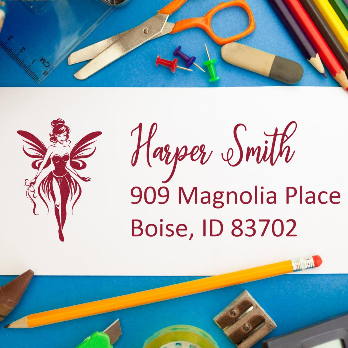 Burgundy custom address stamp and seal with fairy logo for Harper Smith, Boise ID, on white card, surrounded by school supplies.