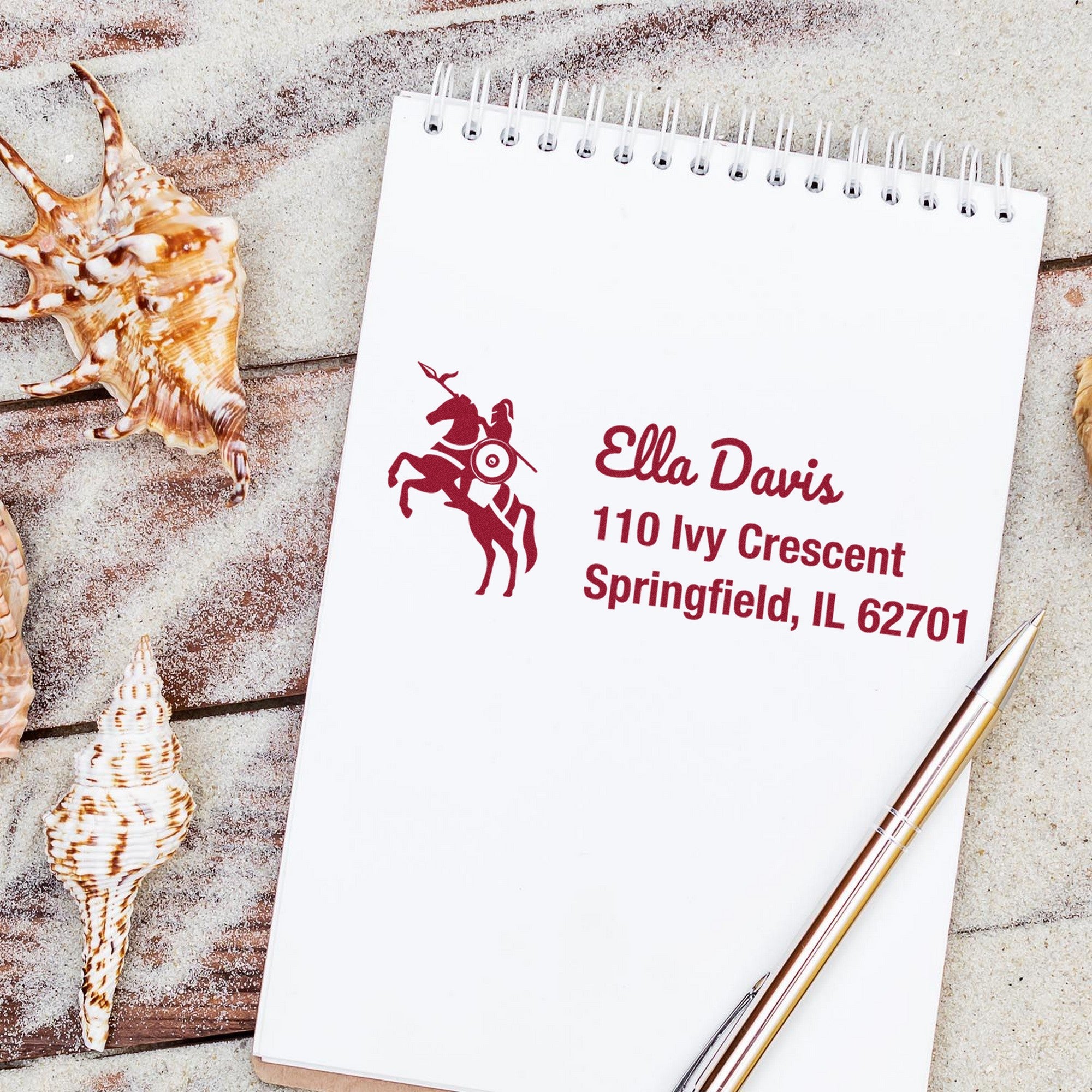 Burgundy custom stamp and seal with horse rider emblem on white notepad, beside seashells and pens.