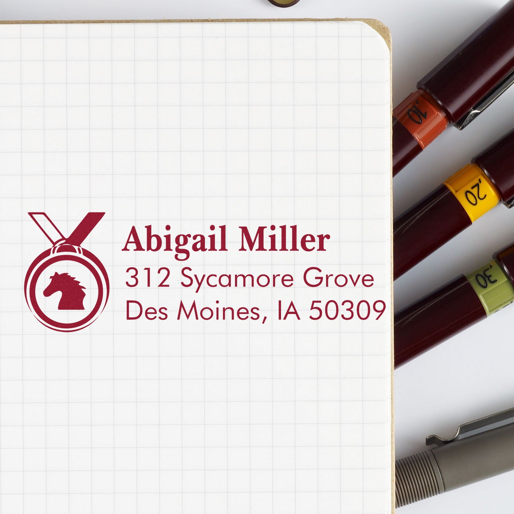 Maroon address stamp and seal on grid paper with horse logo, beside burgundy stamp pens with numbered bands.