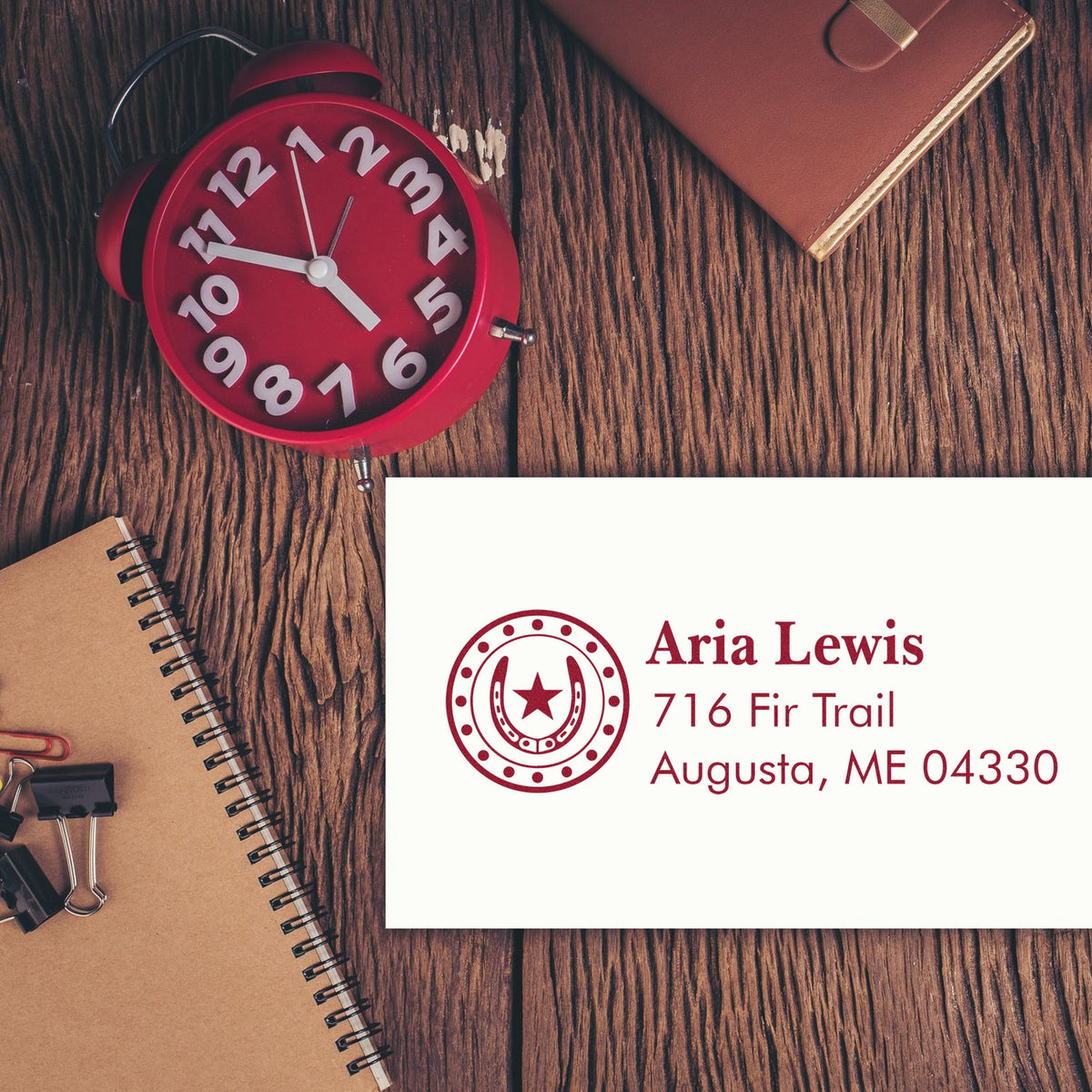 Red custom stamp and seal with address on white card beside a red clock, notebook, and brown leather wallet on wood grain.