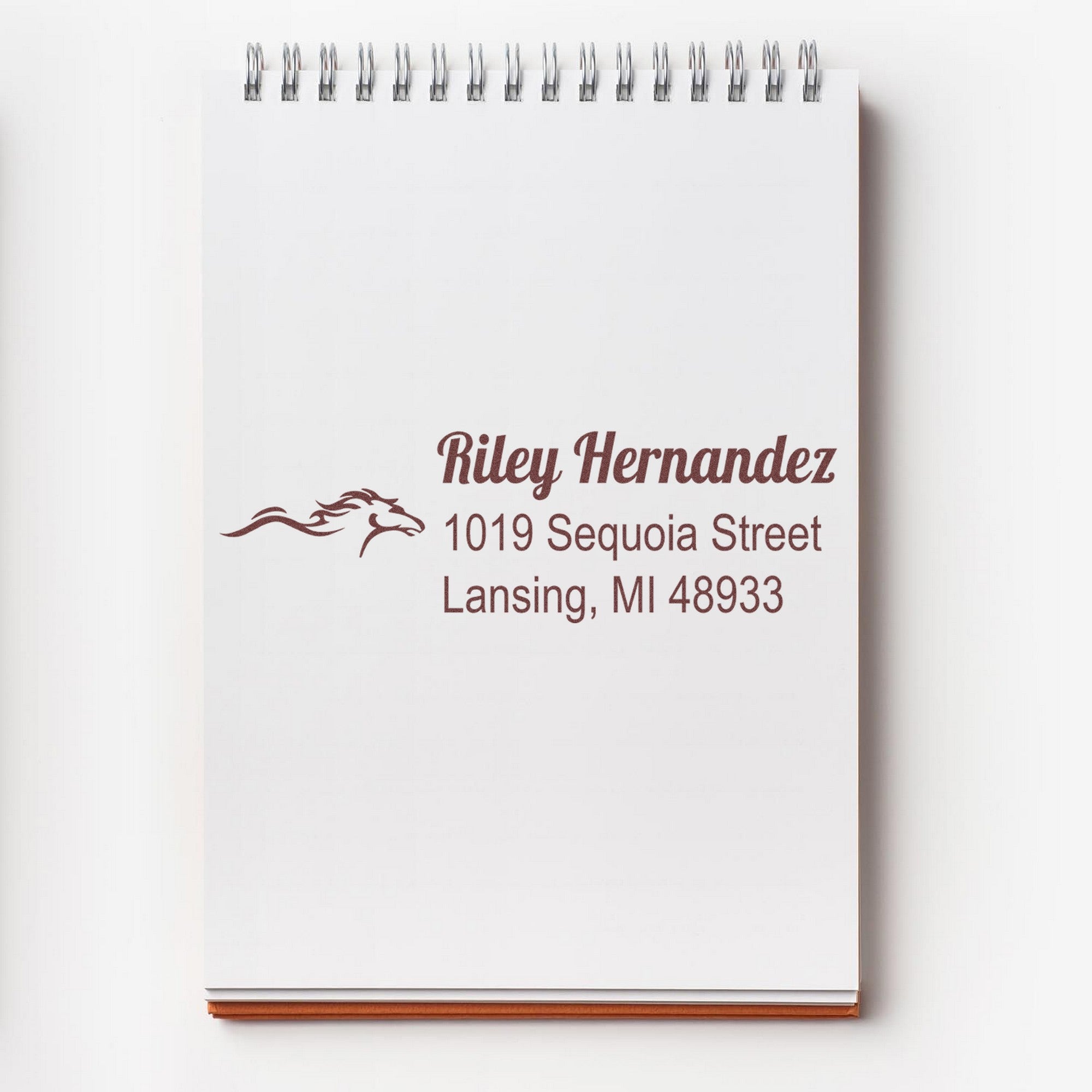 White spiral notepad with burgundy custom address stamp, horse logo, and seal reading Riley Hernandez, Lansing, MI 48933.