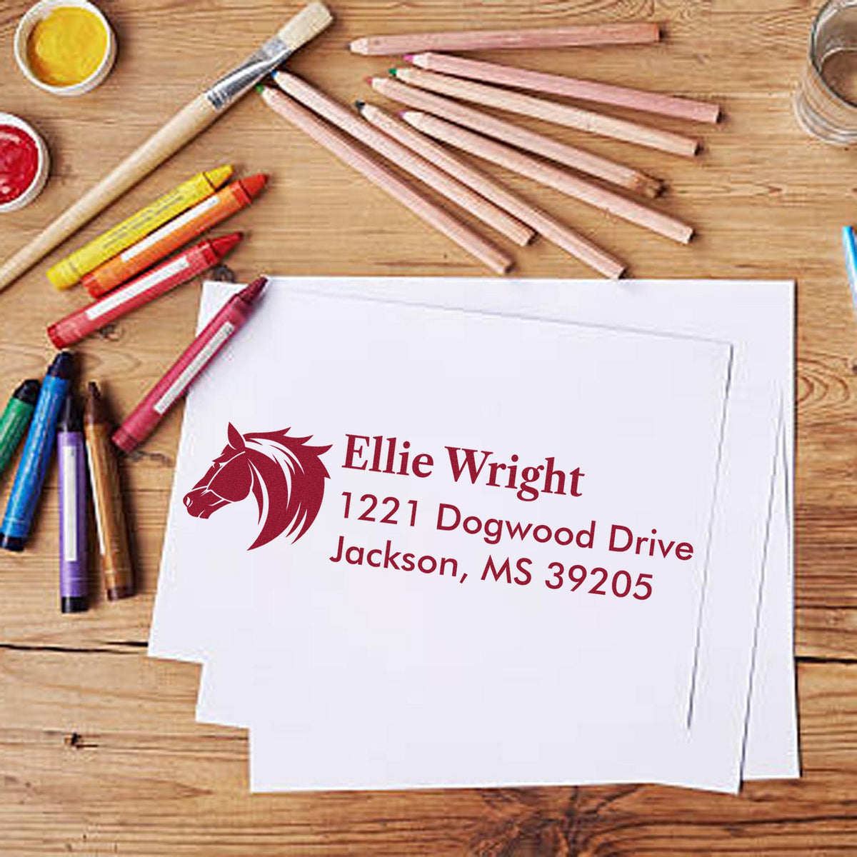 Maroon personalized horse address stamp on white envelope, with pencils and seal tools on a wood desk.
