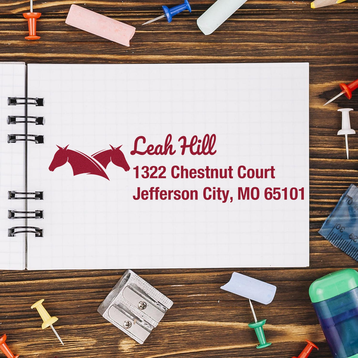 Burgundy custom stamp and seal with horse logo and address on open notebook, surrounded by colorful office supplies.