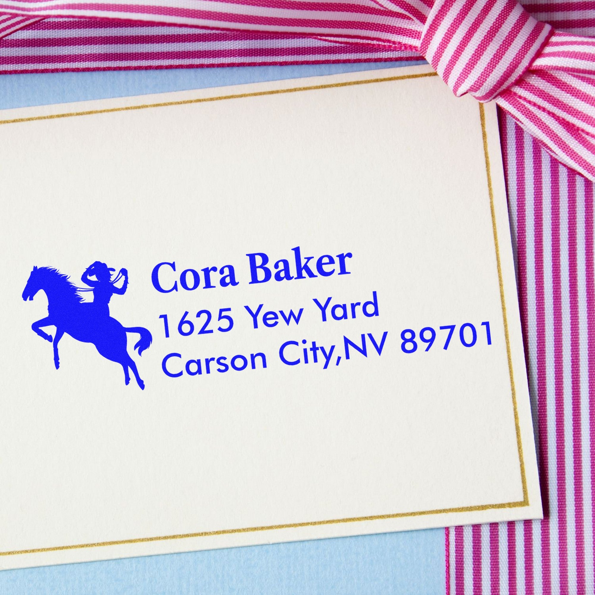 White envelope with blue horse seal and personalized address stamp, accented by pink striped ribbon and gold trim.