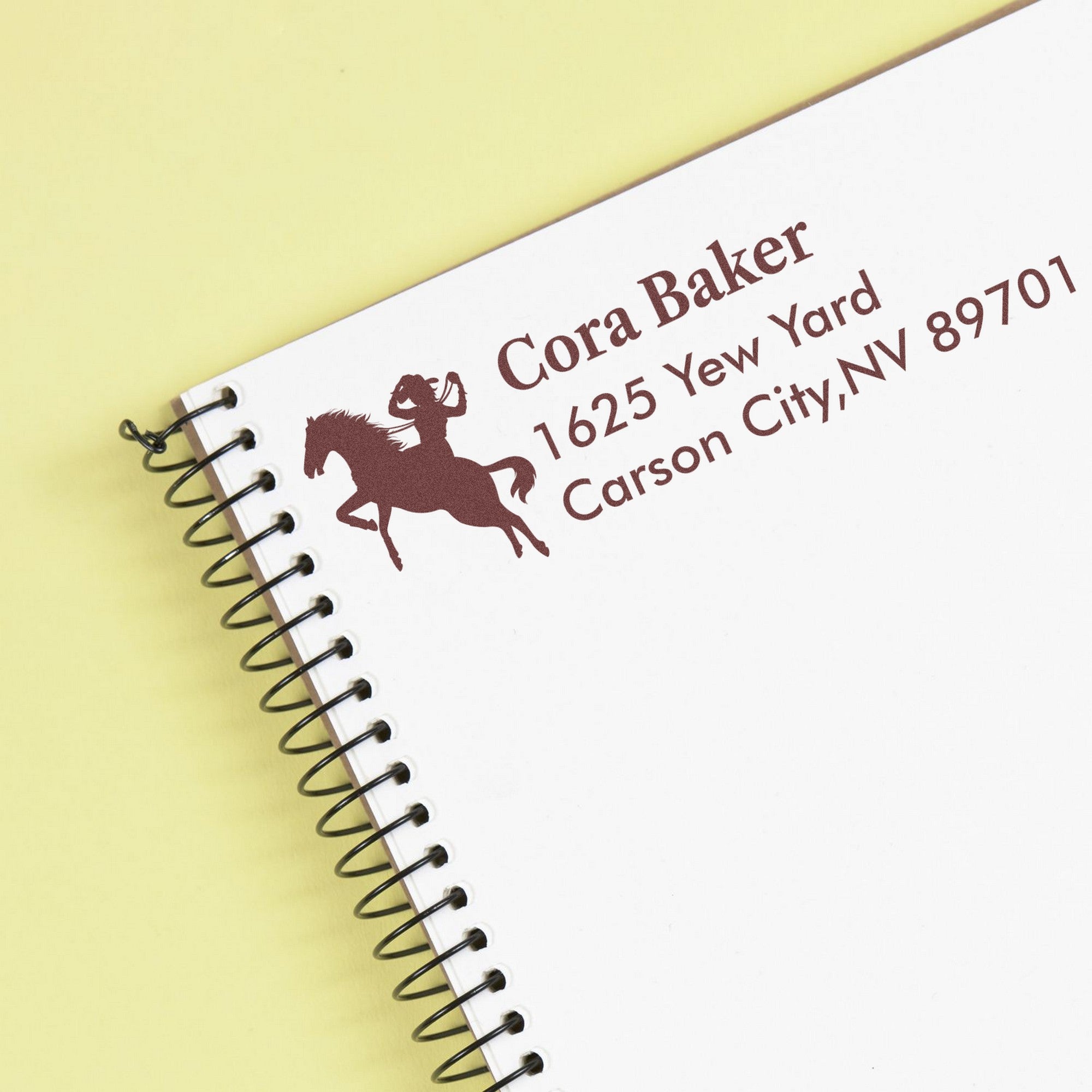 Custom brown horse address stamp and seal with Cora Baker text on a white spiral notebook, pale yellow background