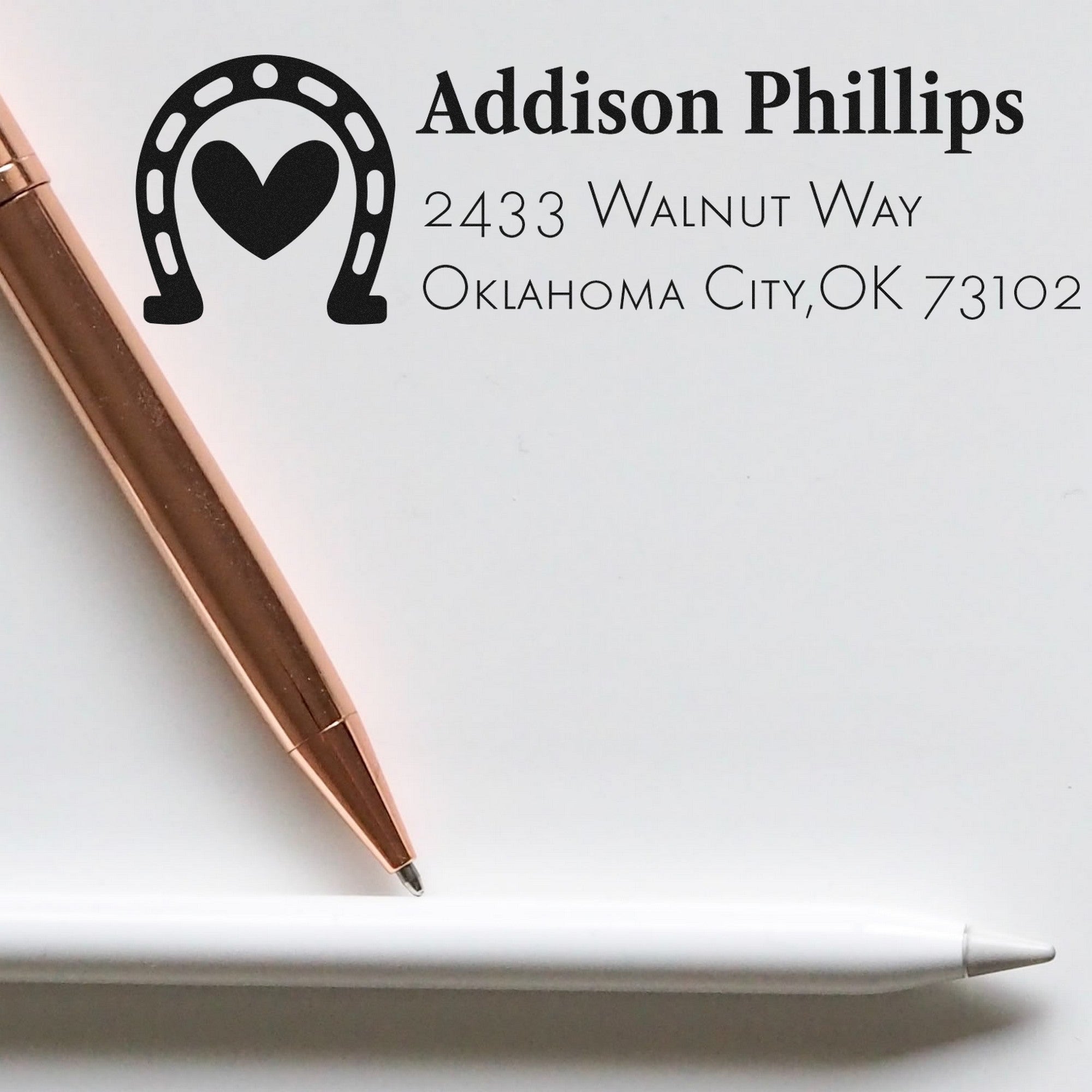 Custom black address stamp and seal with horseshoe heart logo, Addison Phillips text, on white paper with rose gold tool.