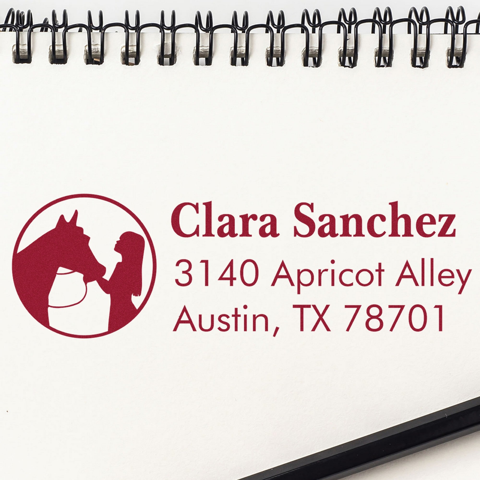 Maroon custom address stamp with horse-and-woman seal, Clara Sanchez, 3140 Apricot Alley, Austin TX 78701 on white spiral notebook