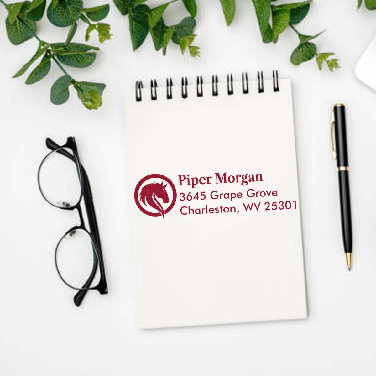 Custom burgundy address stamp and seal on white spiral notebook, with black glasses, pen, and green leaves.