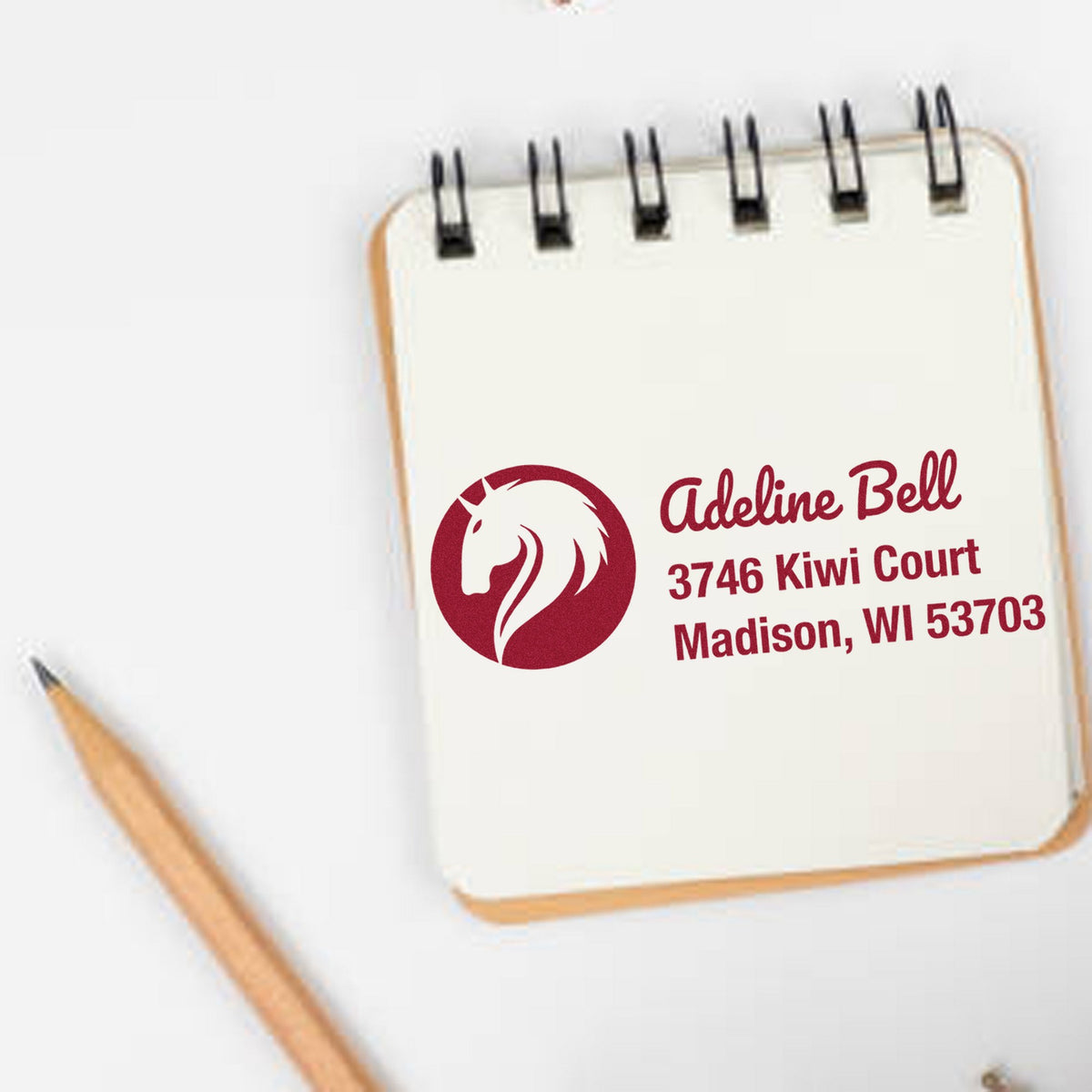Custom red address stamp and seal with horse logo on cream spiral notepad, beside a pencil.