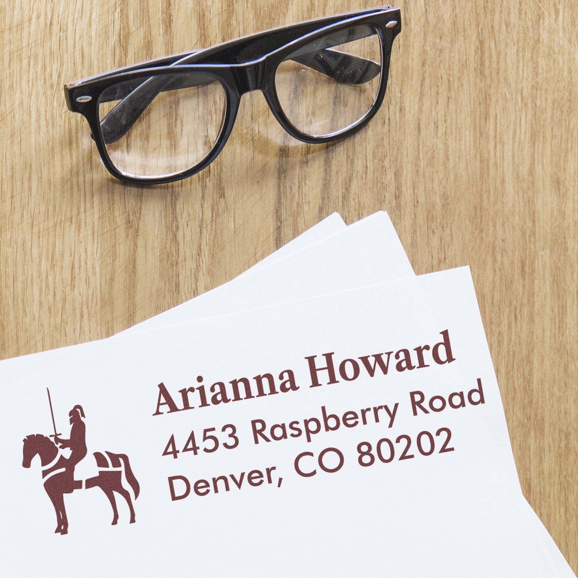 Black eyeglasses next to a brown custom address stamp and seal on white paper over a wood desk.