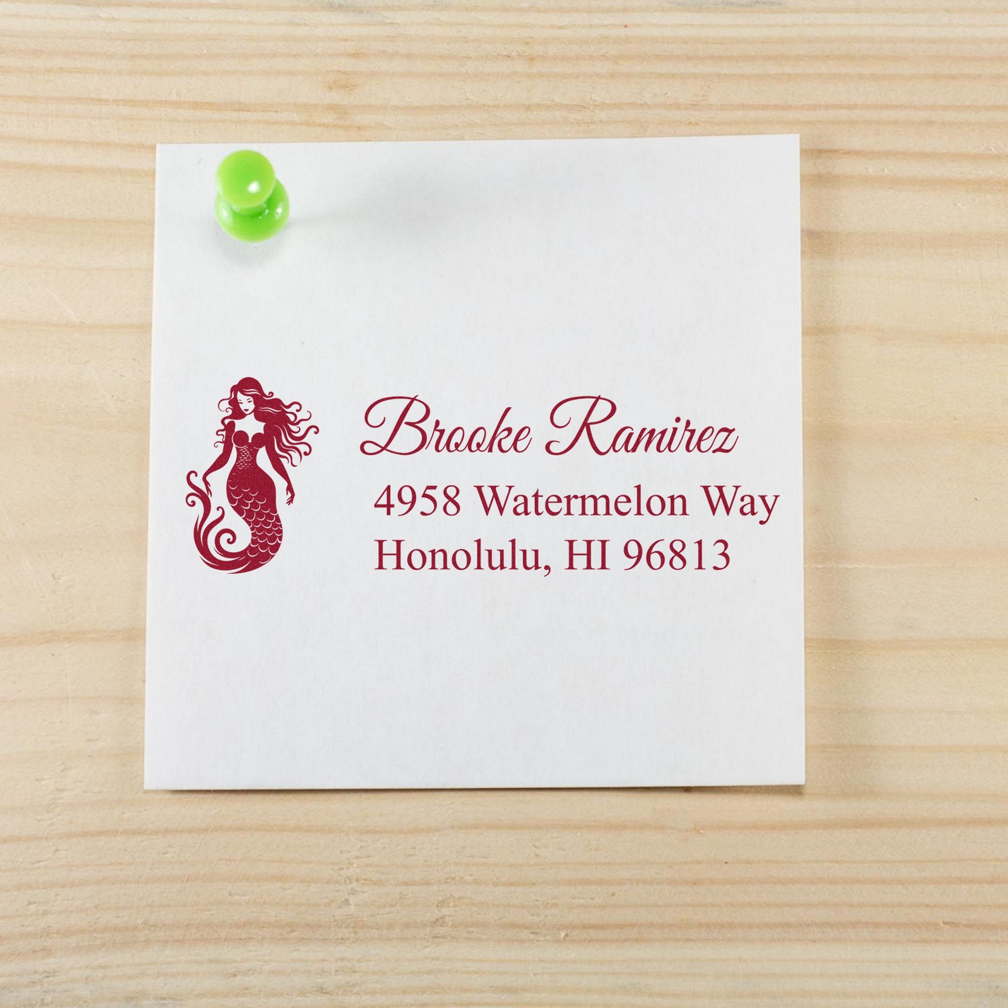 Custom stamp and seal sample on white card with maroon mermaid logo and address, pinned with a green pushpin on wood.