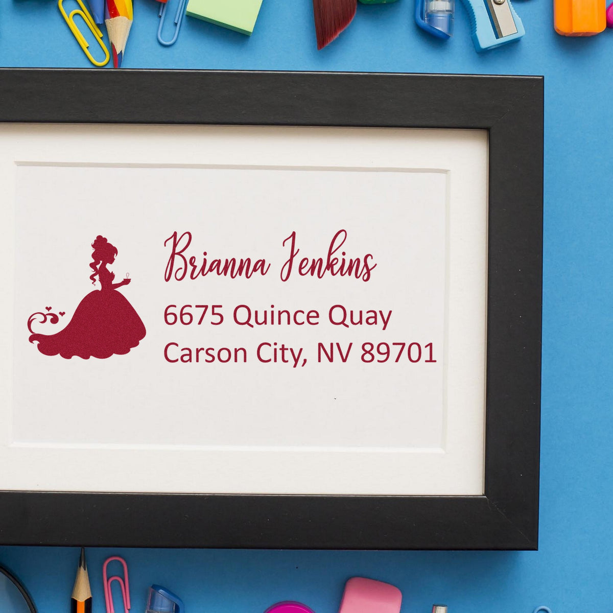 Burgundy princess address stamp seal in black frame on white paper, with blue background and colorful office supplies.
