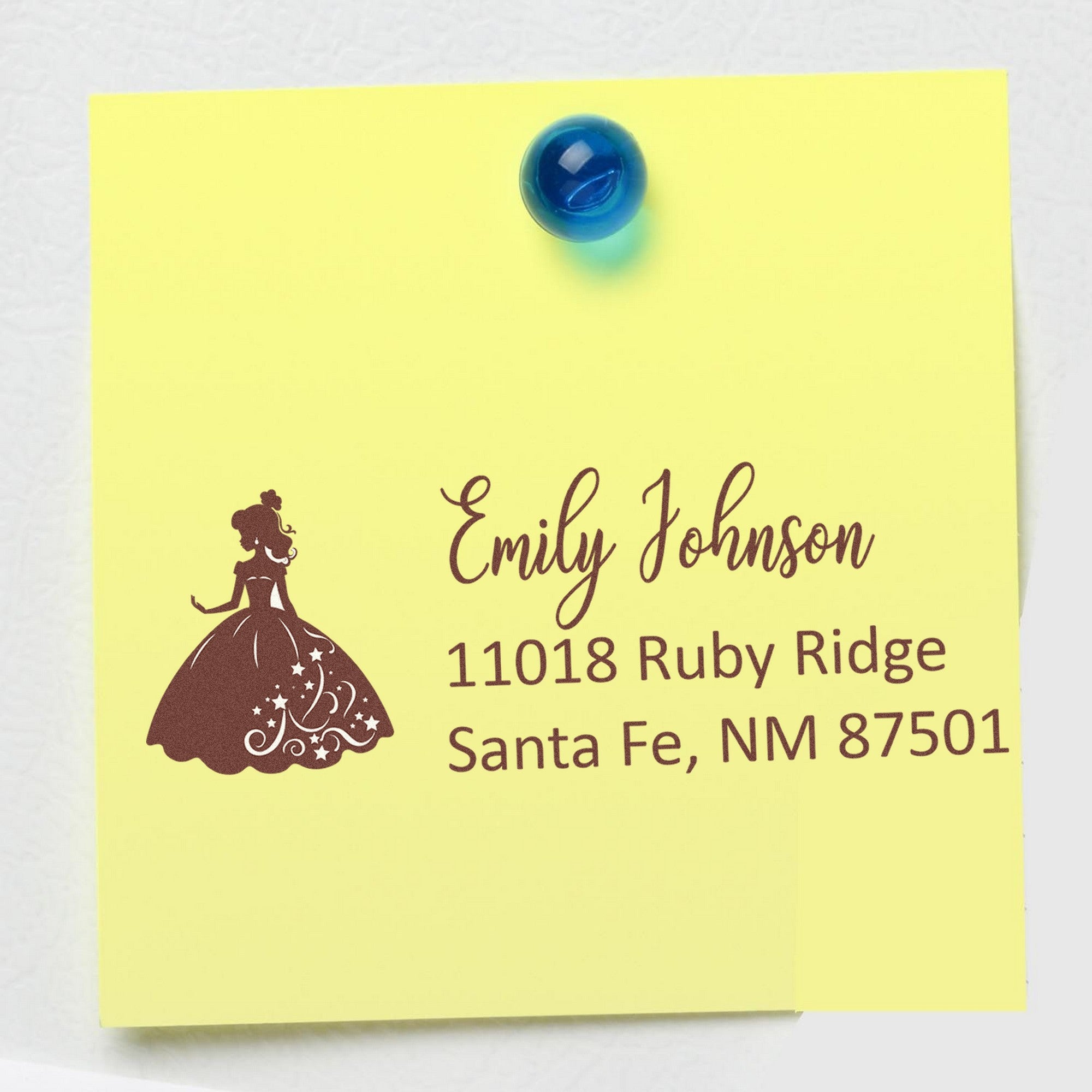 Yellow note pinned with a blue tack, featuring a brown custom address stamp with princess silhouette, Emily Johnson, Santa Fe NM 87501.