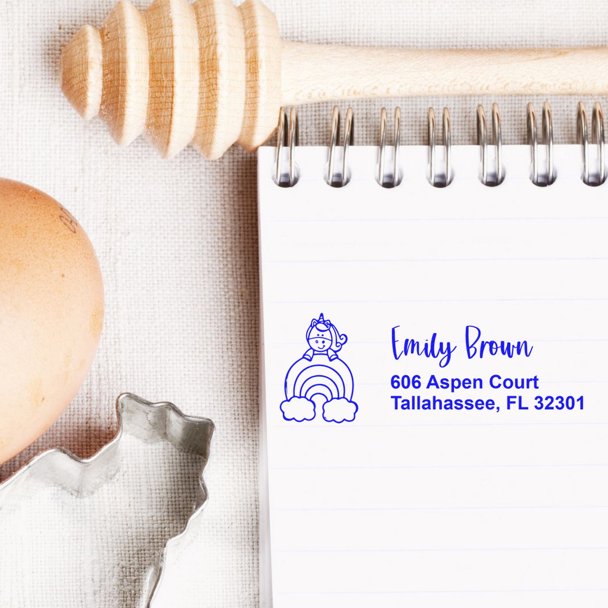 Blue personalized stamp seal on notepad with unicorn rainbow design, Emily Brown name and address, beside wooden rolling pin and cookie cutters.