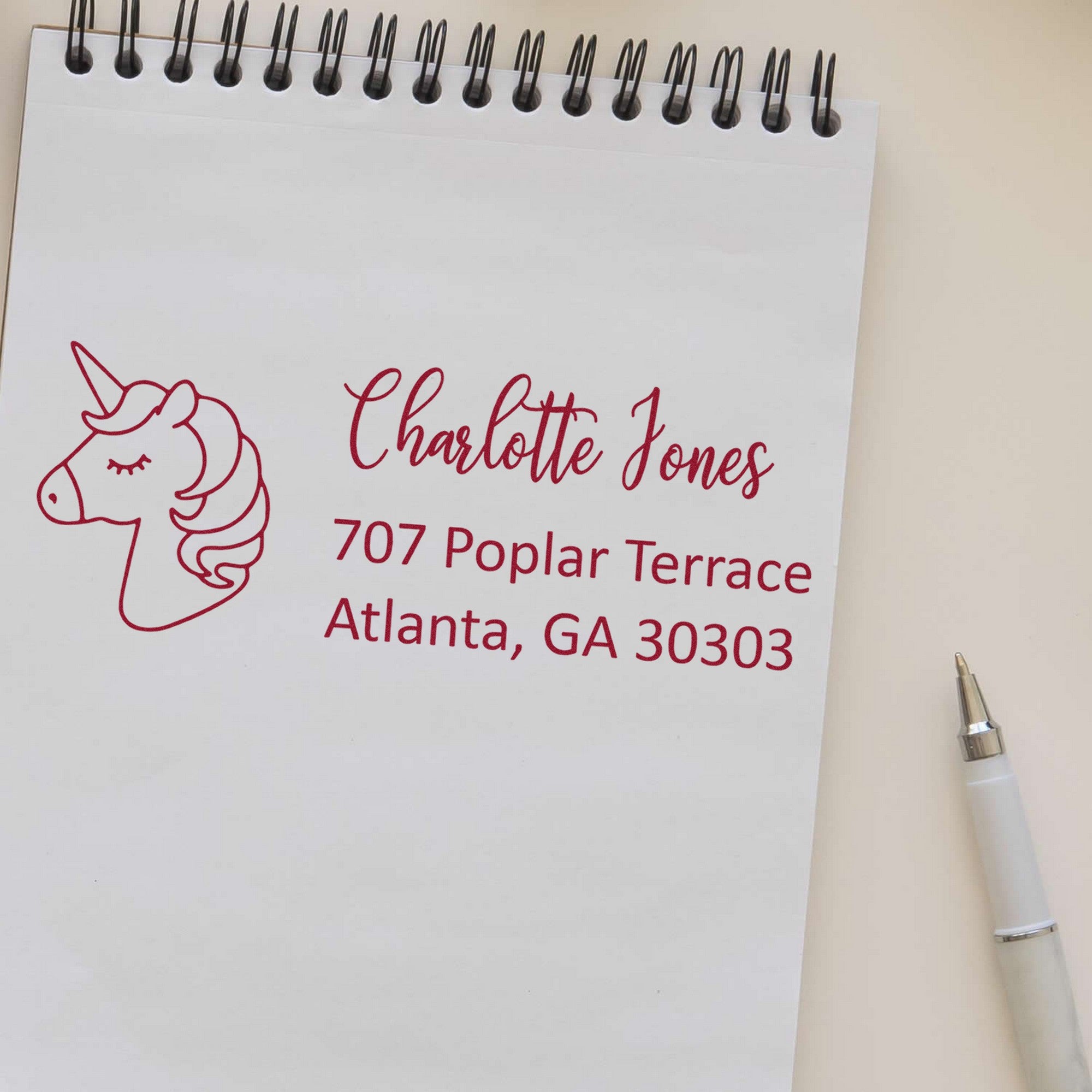 Magenta custom address stamp with unicorn seal on white spiral notepad, reading Charlotte Jones, 707 Poplar Terrace, Atlanta, GA 30303.