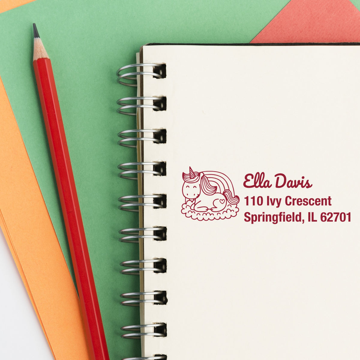 Custom unicorn address stamp on cream spiral notebook, beside a red pencil with green and orange paper background.