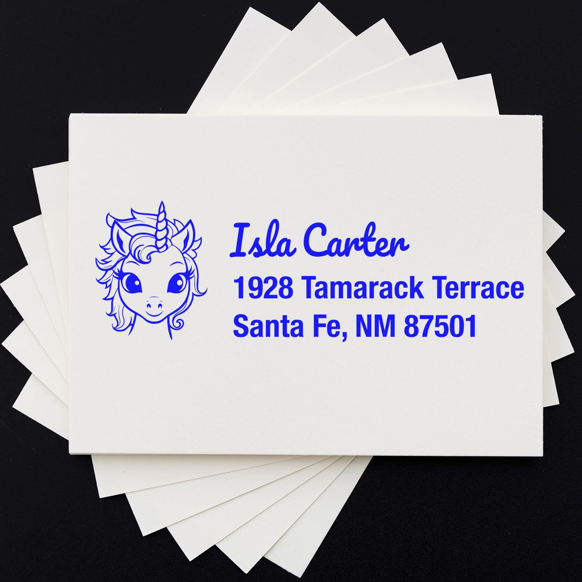 White stamp and seal cards with blue unicorn logo and address for Isla Carter, Santa Fe, NM, on a black background.