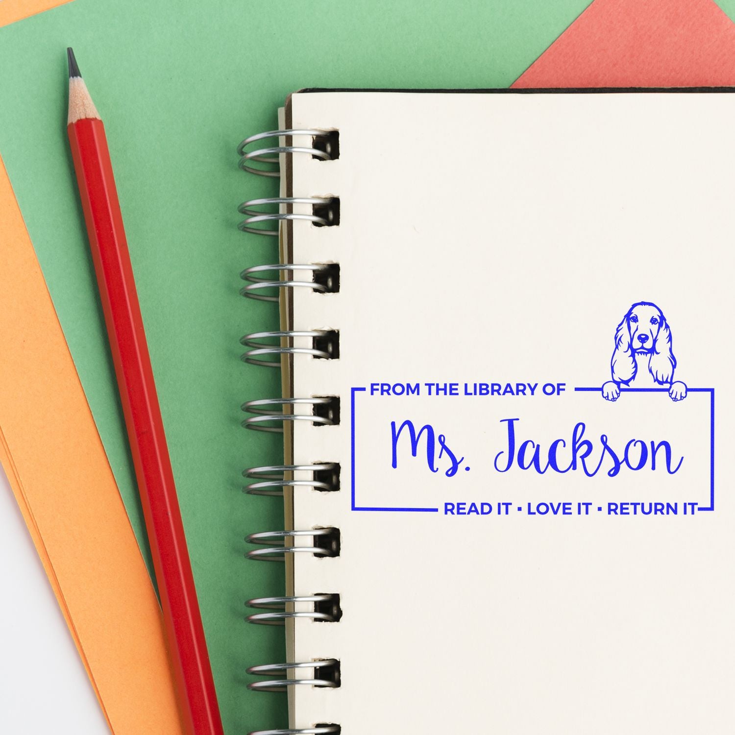 Custom blue library stamp reading Ms. Jackson on a spiral notebook beside a red pencil and colorful paper.