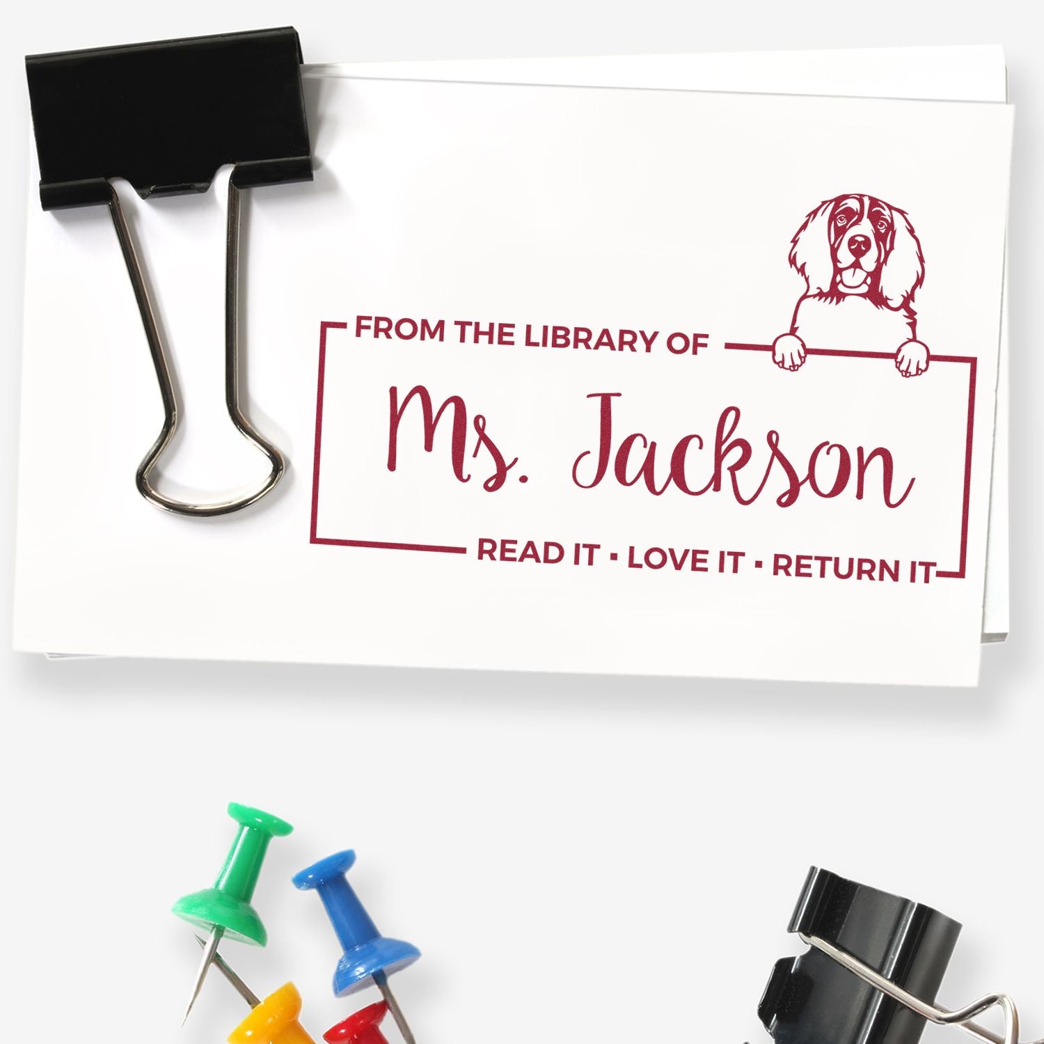Custom burgundy library stamp reading Ms. Jackson with dog graphic, clipped to white card; black binder clip and colorful pushpins.