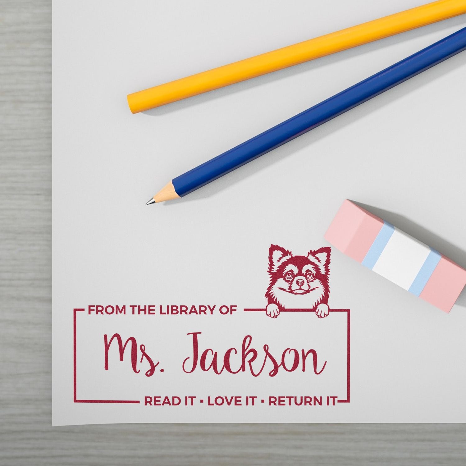 Red custom library stamp and seal with dog mascot on white paper, beside yellow and blue pencils and a pink eraser.