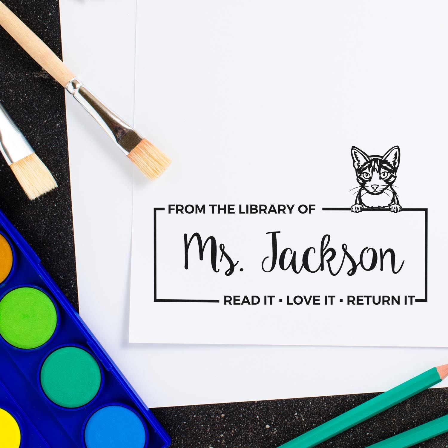 Custom library stamp and seal with cat outline, black text on white, surrounded by paintbrushes and colorful art supplies.