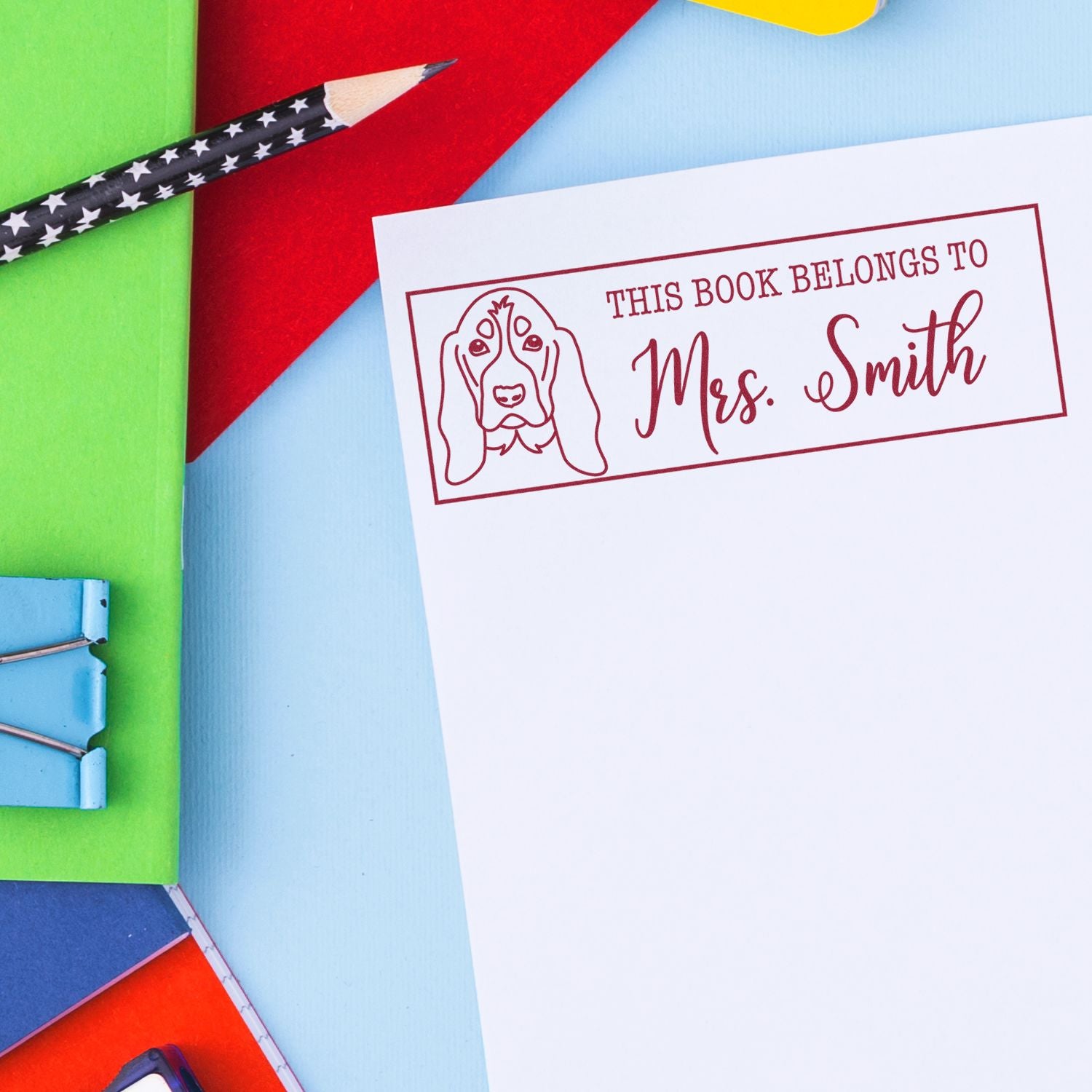 Red custom book stamp with dog illustration and Mrs. Smith on white paper, against a colorful stationery background.