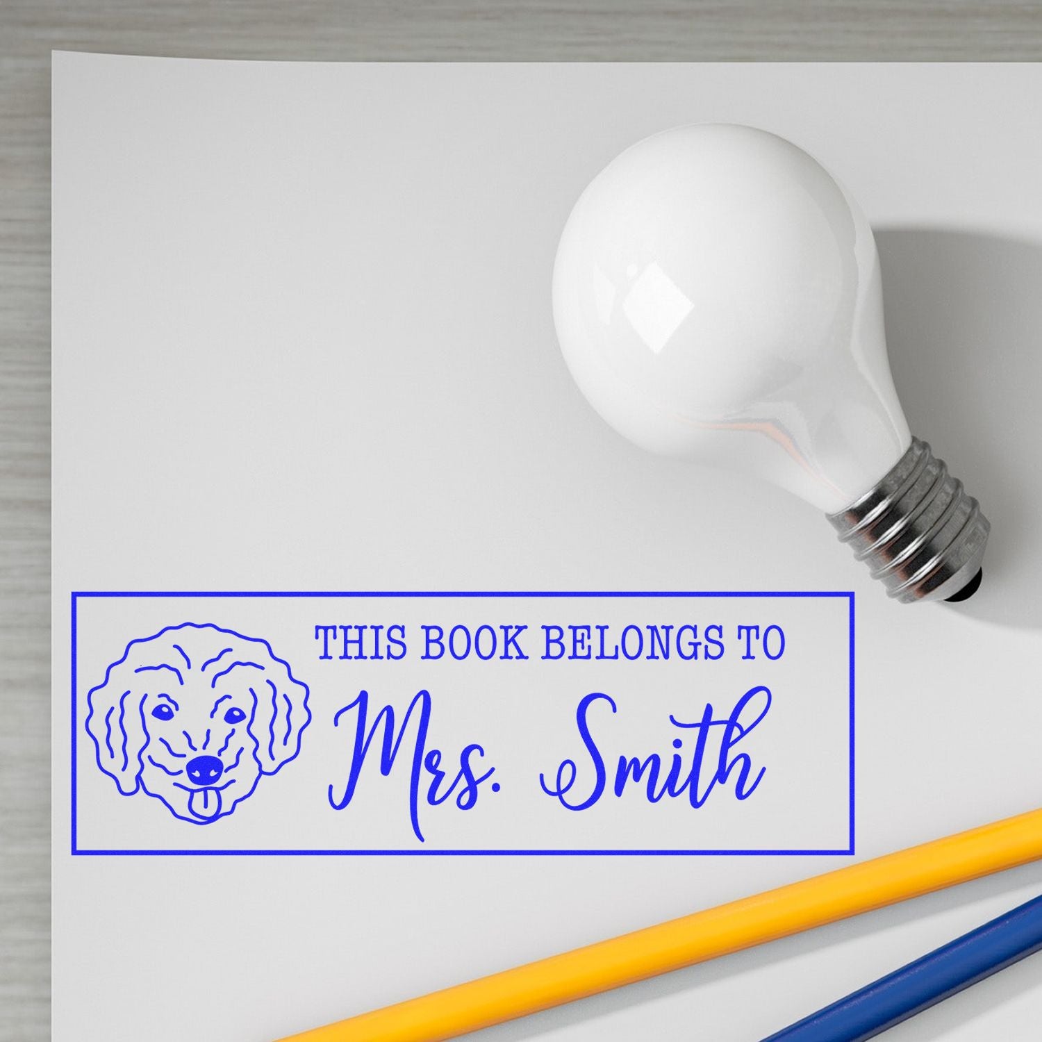 White lightbulb-shaped stamp/seal above blue bookplate label with dog icon and This Book Belongs to Mrs. Smith; yellow and blue pencils