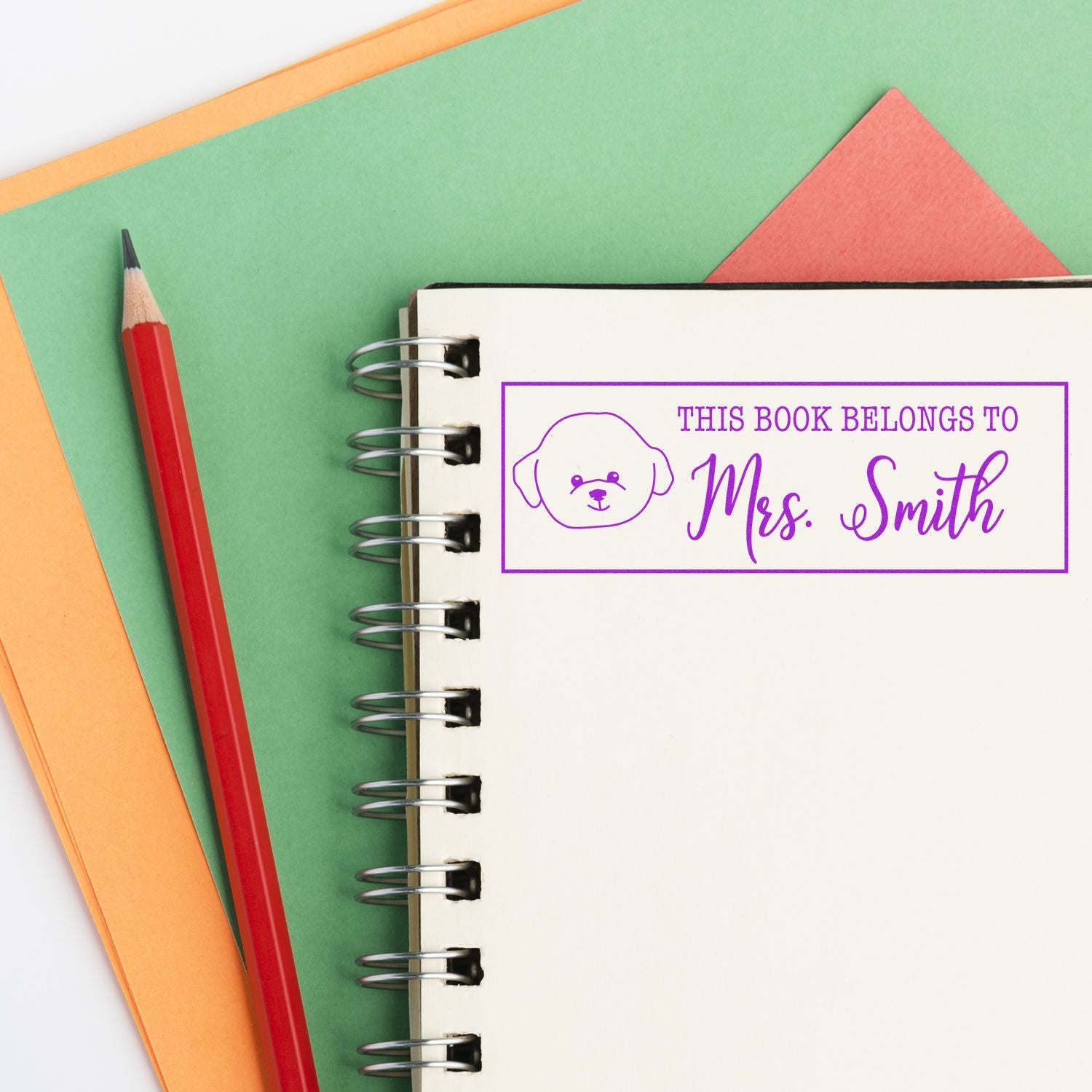 Purple custom book ownership stamp seal with dog icon on a spiral notebook, beside a red pencil on green and orange paper.