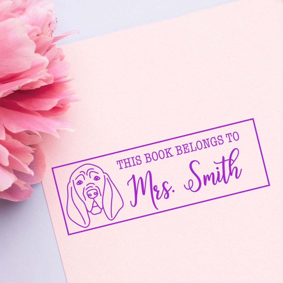 Purple custom book ownership stamp and seal with dog outline, reading This Book Belongs to Mrs. Smith on soft pink paper.